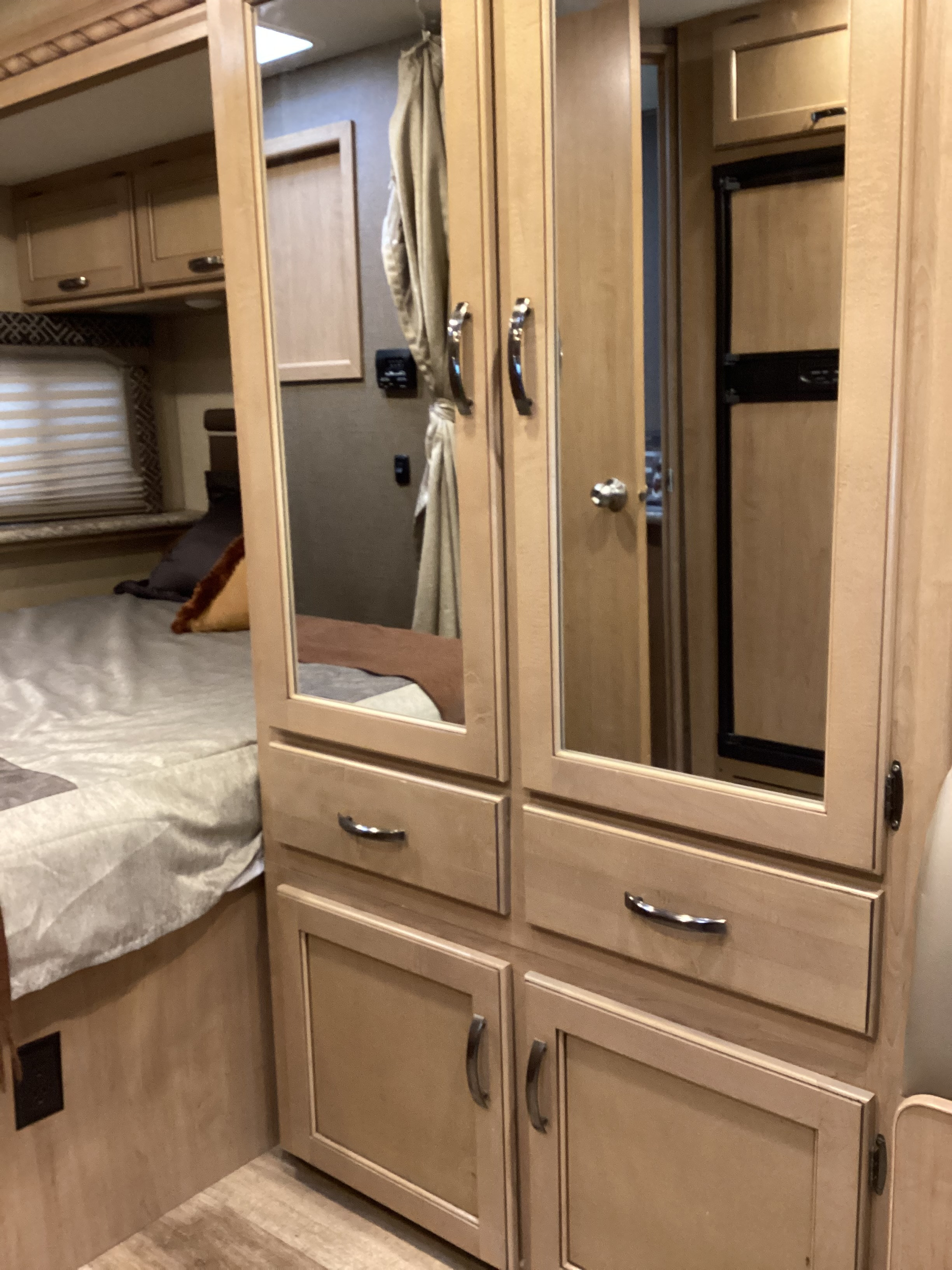 Cozy RV rental bedroom featuring a made bed, window with blinds, and mirrored wooden cabinetry reflecting the interior and a fridge.. Thor Four Winds 2018