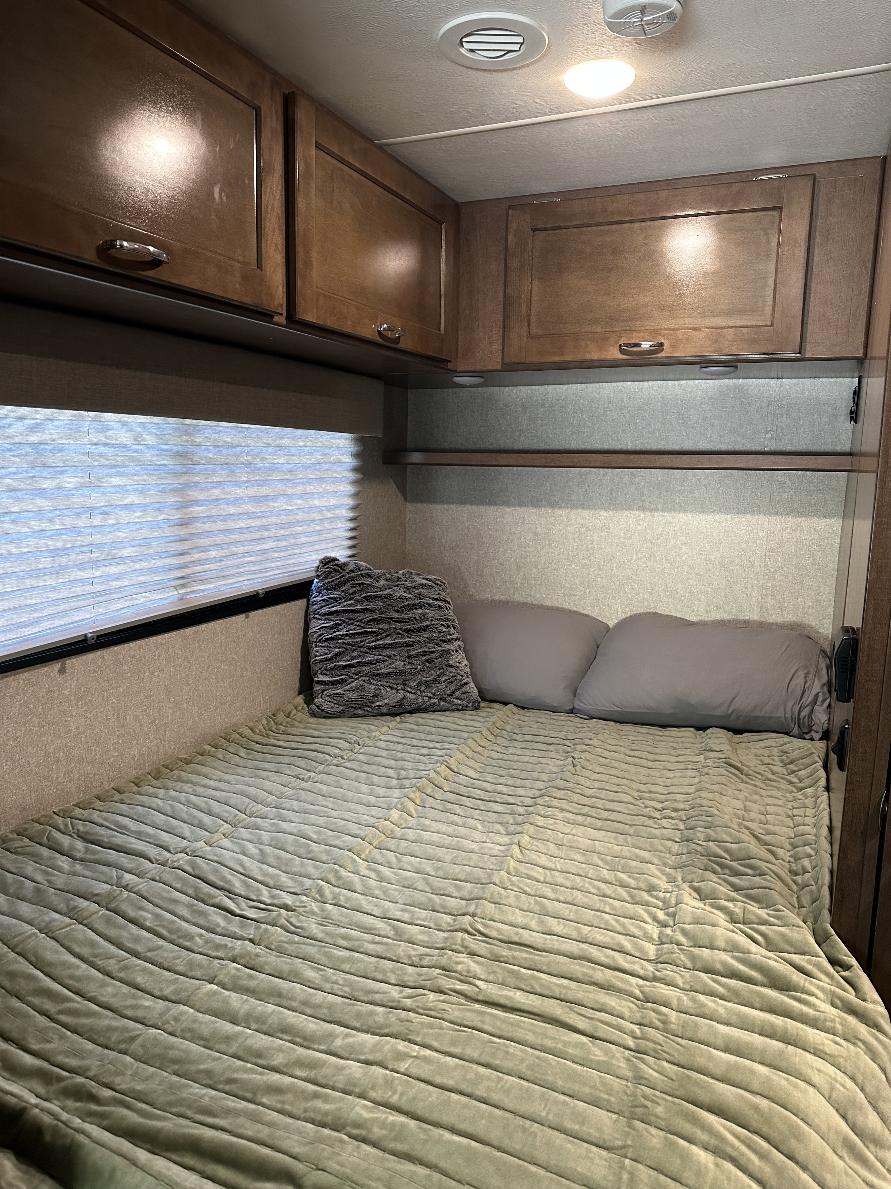 Cozy RV rental bedroom featuring a plush, ribbed sage green comforter, gray pillows, and overhead wood storage cabinets.. Winnebago Vita 2020