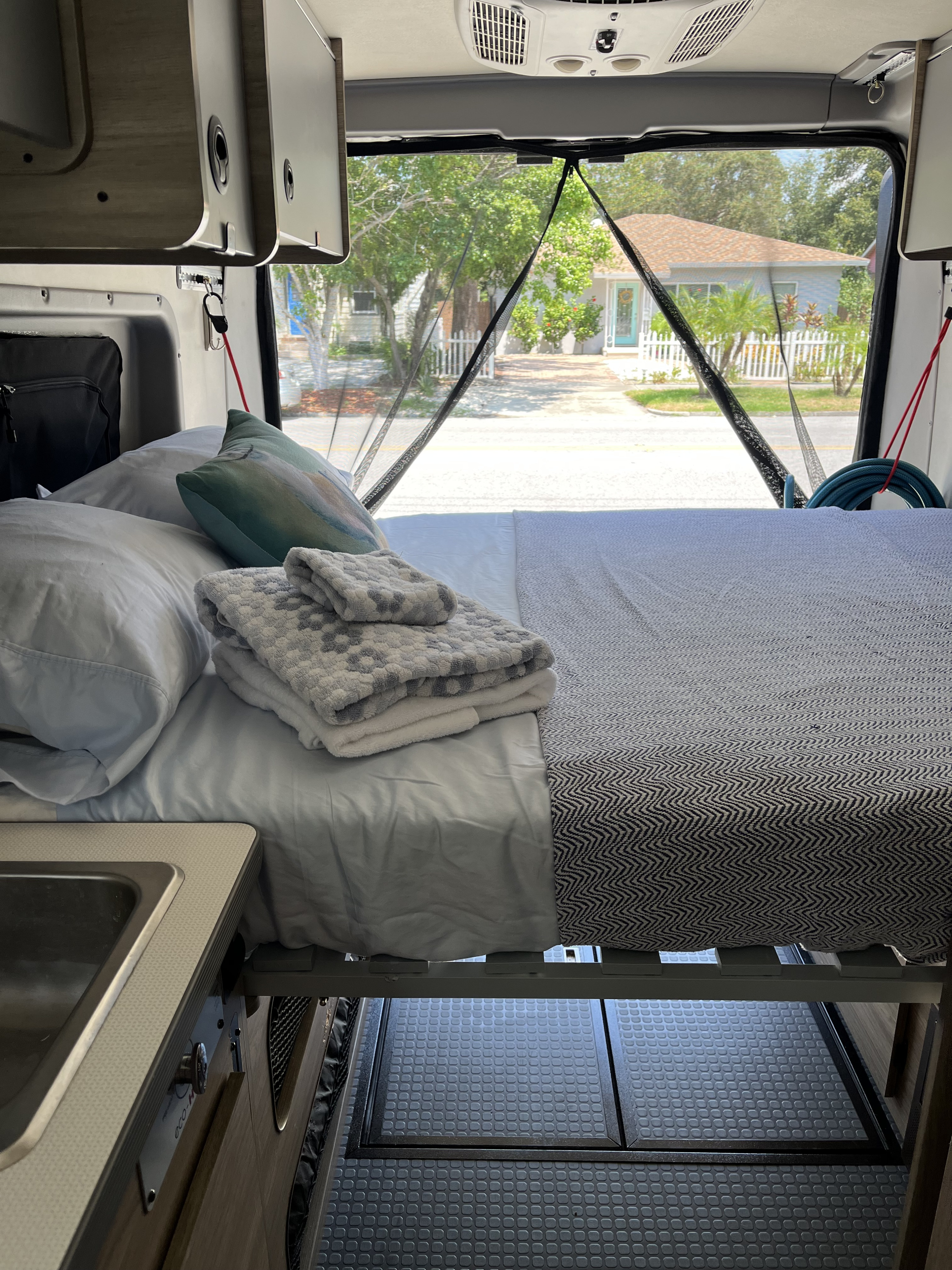 RV rental interior featuring a cozy bed, kitchenette, and a view of a charming street through the mesh screen.. Winnebago Solis 59PX 2021