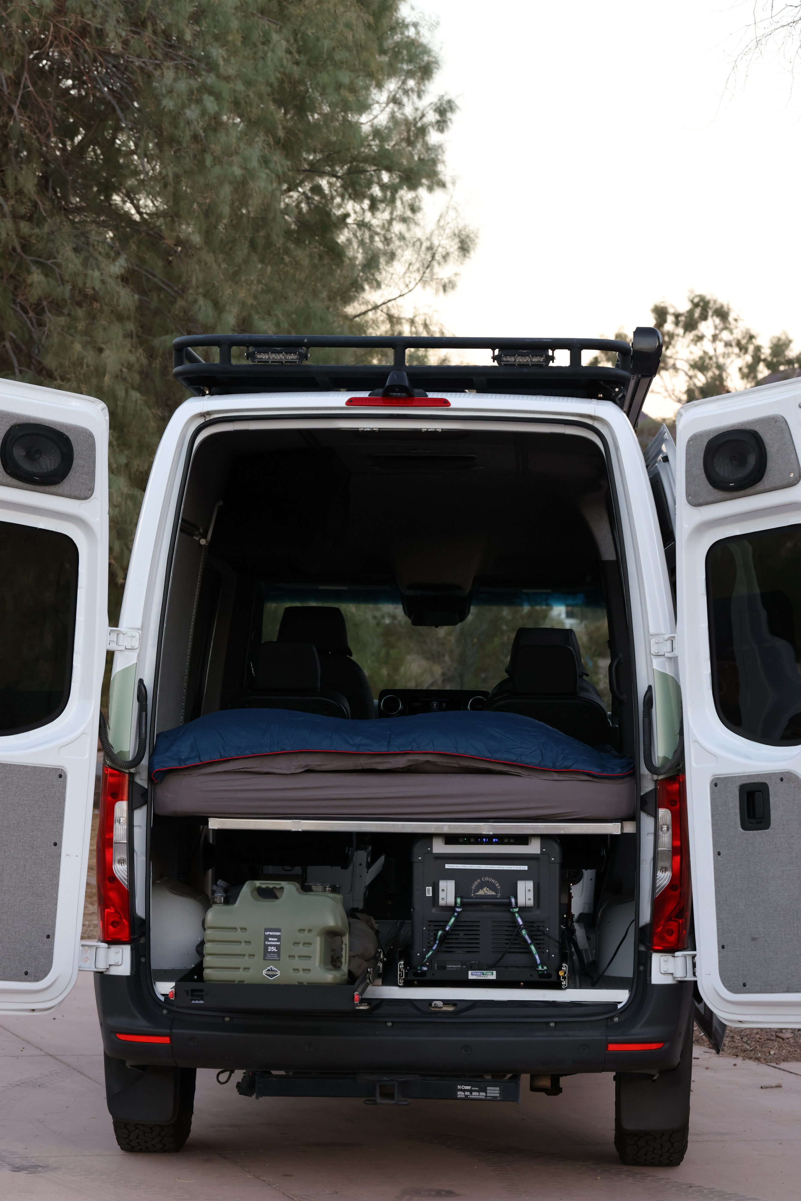 Explore this spacious RV rental camper van with open rear doors, revealing a comfortable bed, portable fridge, and water storage.. Mercedes Benz Sprinter 2019