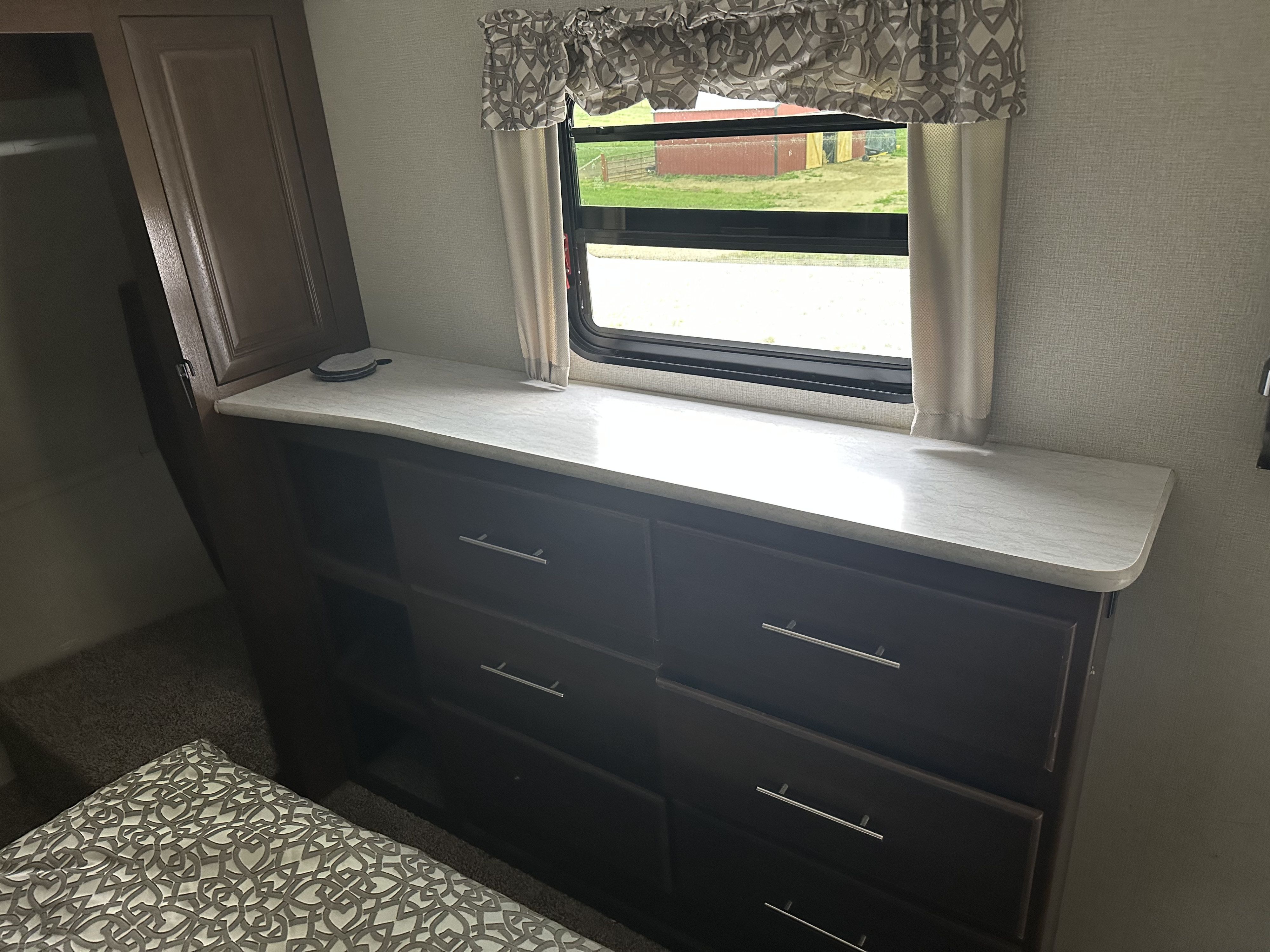 This RV rental bedroom features a dark wood dresser, a window with patterned valance, and part of a bed, offering practical storage.. Forest River Wildcat 2018