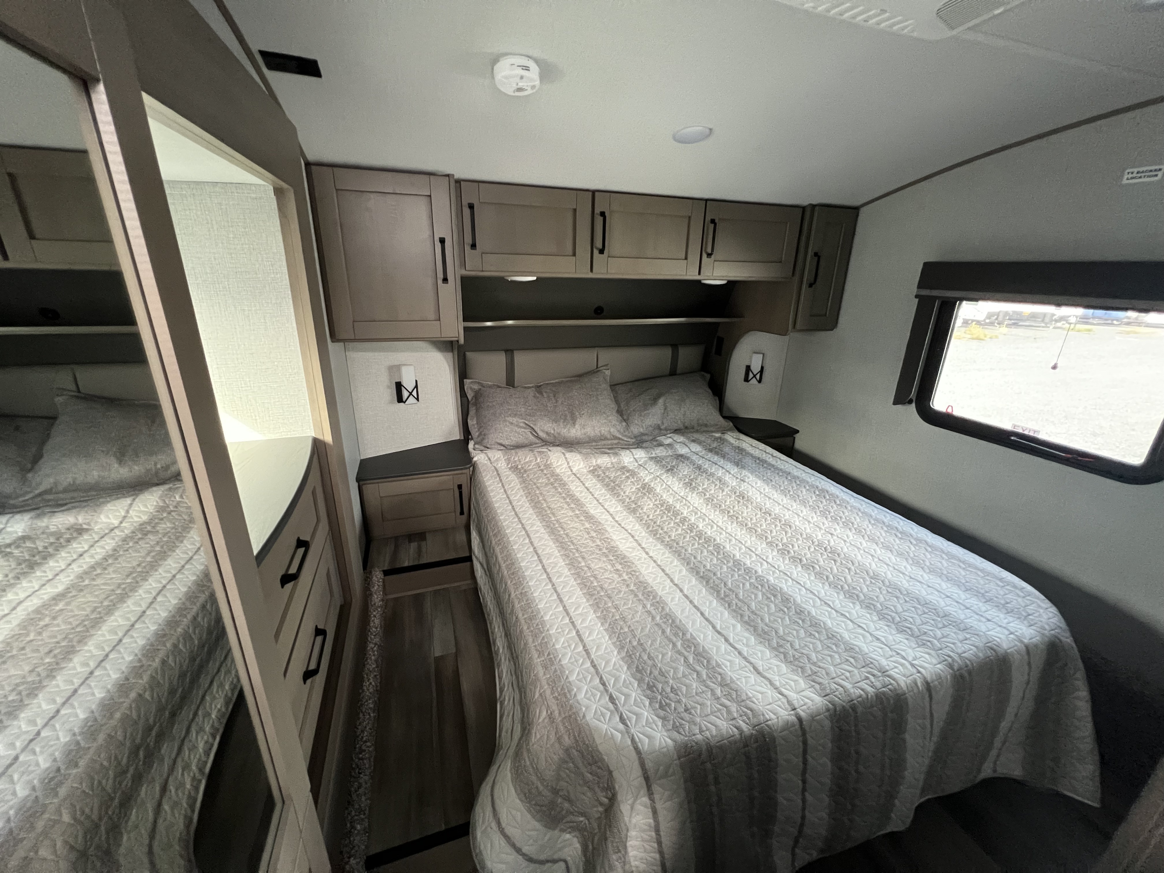 Inside a modern RV rental, this cozy bedroom offers a queen bed, overhead cabinets, a mirrored wardrobe, and a window.. Grand Design Reflection 2023