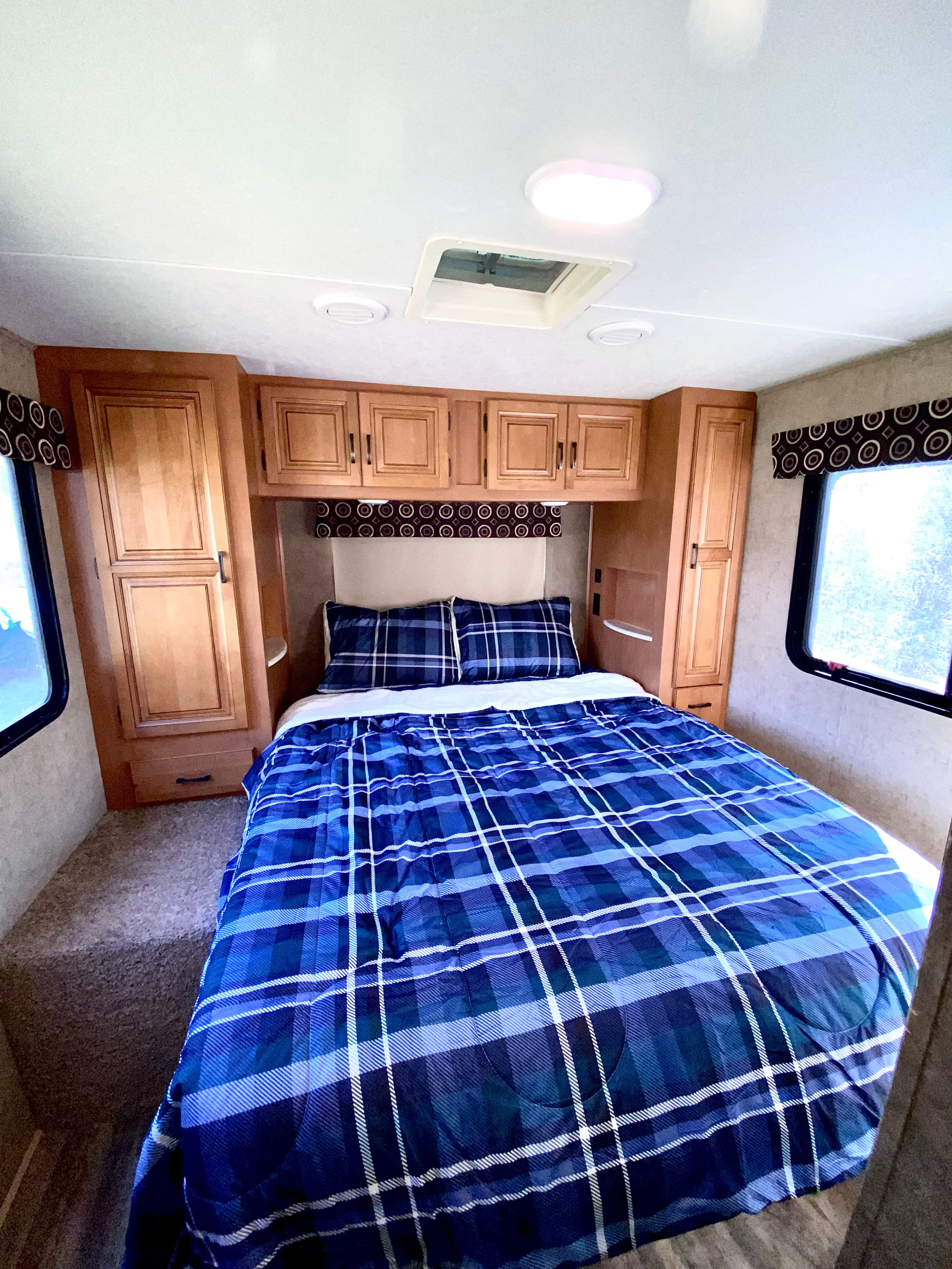 Cozy RV rental bedroom features a comfy bed with blue plaid bedding, wood cabinets, and bright windows.. Forest River Sunseeker 2016