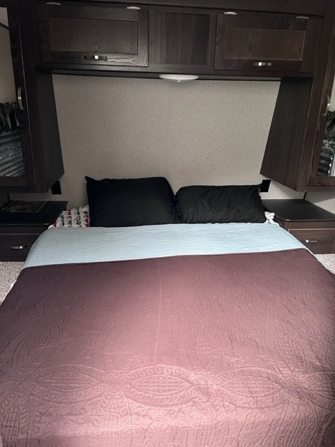 This RV rental bedroom features a comfy bed with black pillows, maroon quilted blanket, and dark wood built-in storage.. Keystone RV Springdale 2019