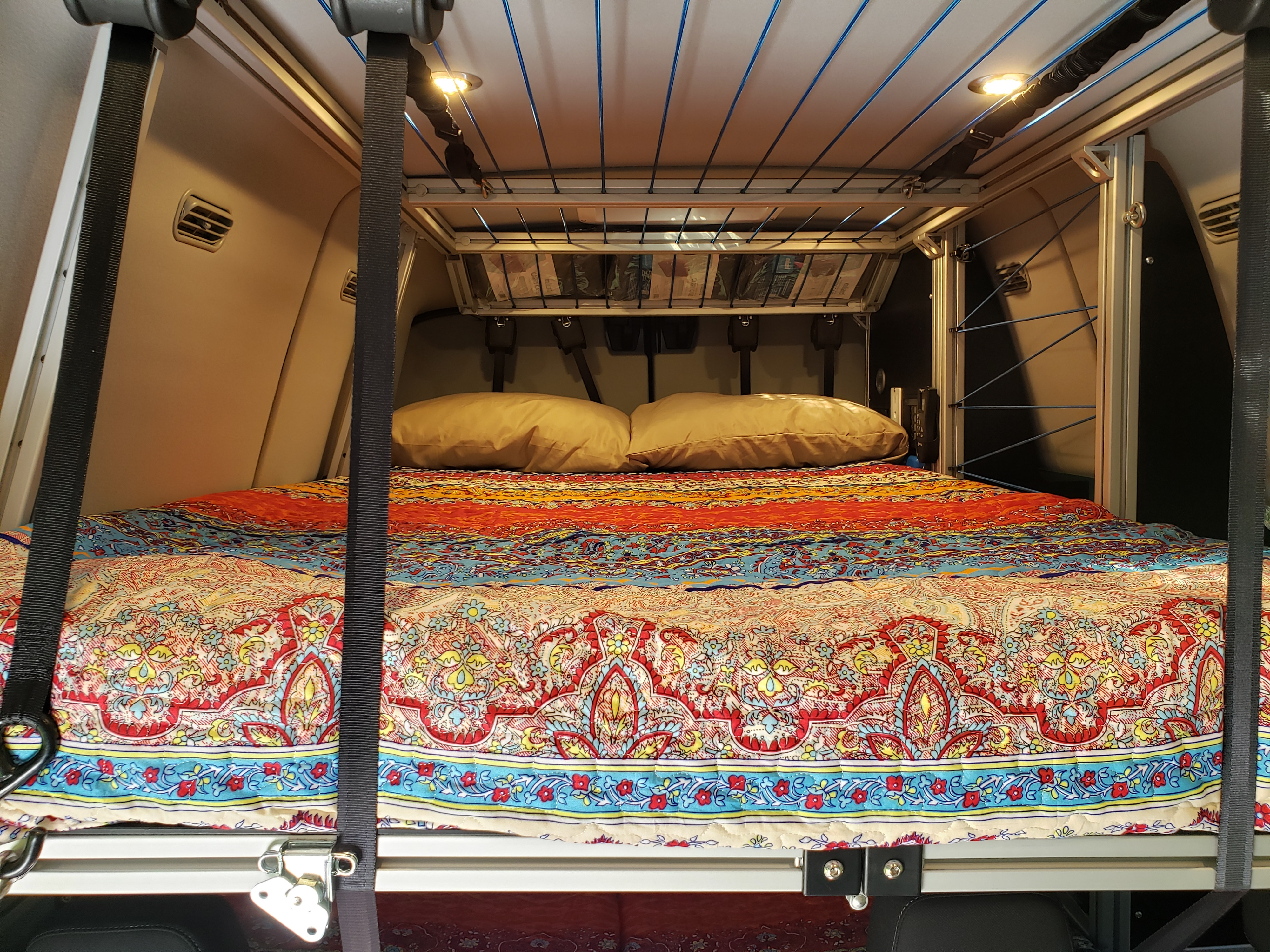 Comfortable bed inside an RV rental, featuring a vibrant patterned comforter, pillows, and custom overhead storage.. Ford Transit 2022