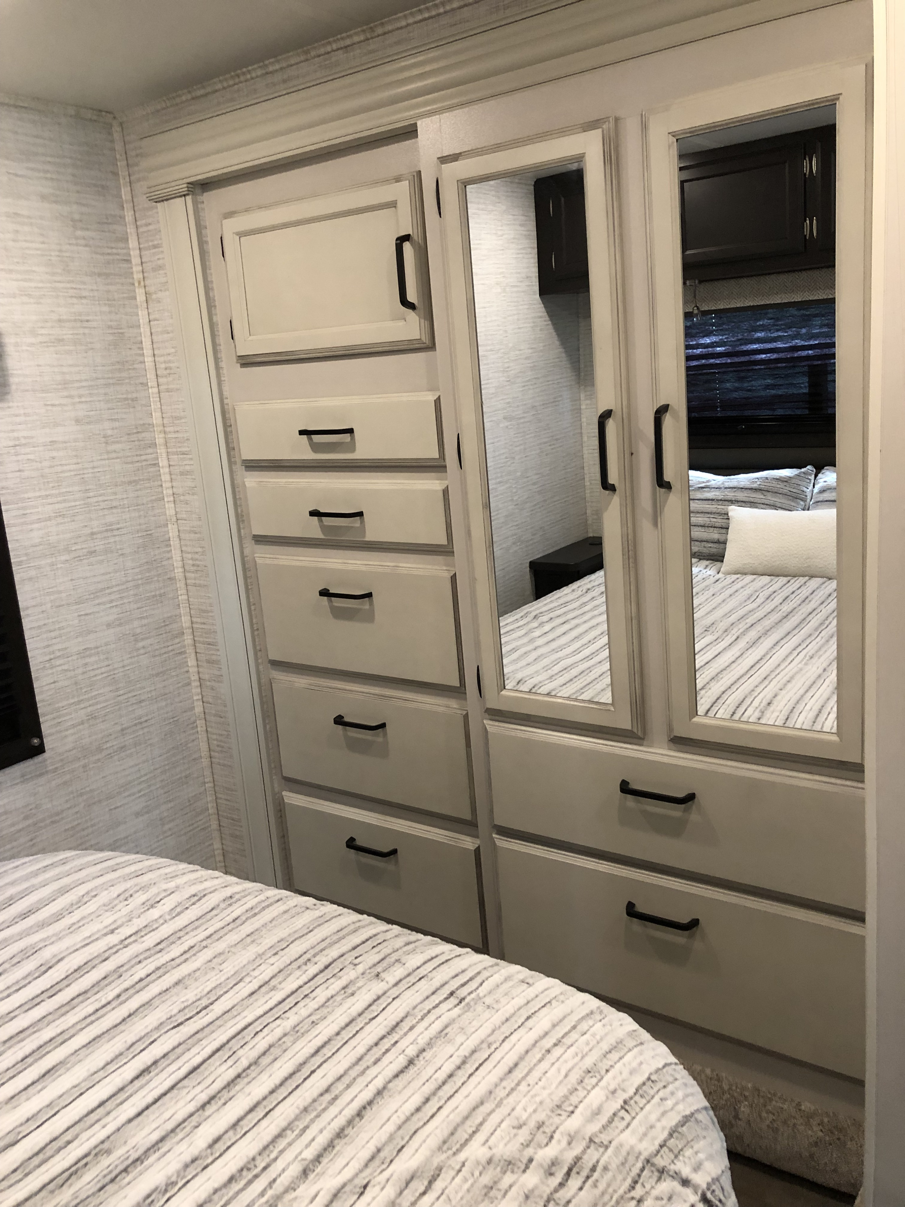 RV rental bedroom with modern light gray dresser/wardrobe, featuring mirrored doors reflecting the striped bed. Functional storage.. Jayco Redhawk 2024