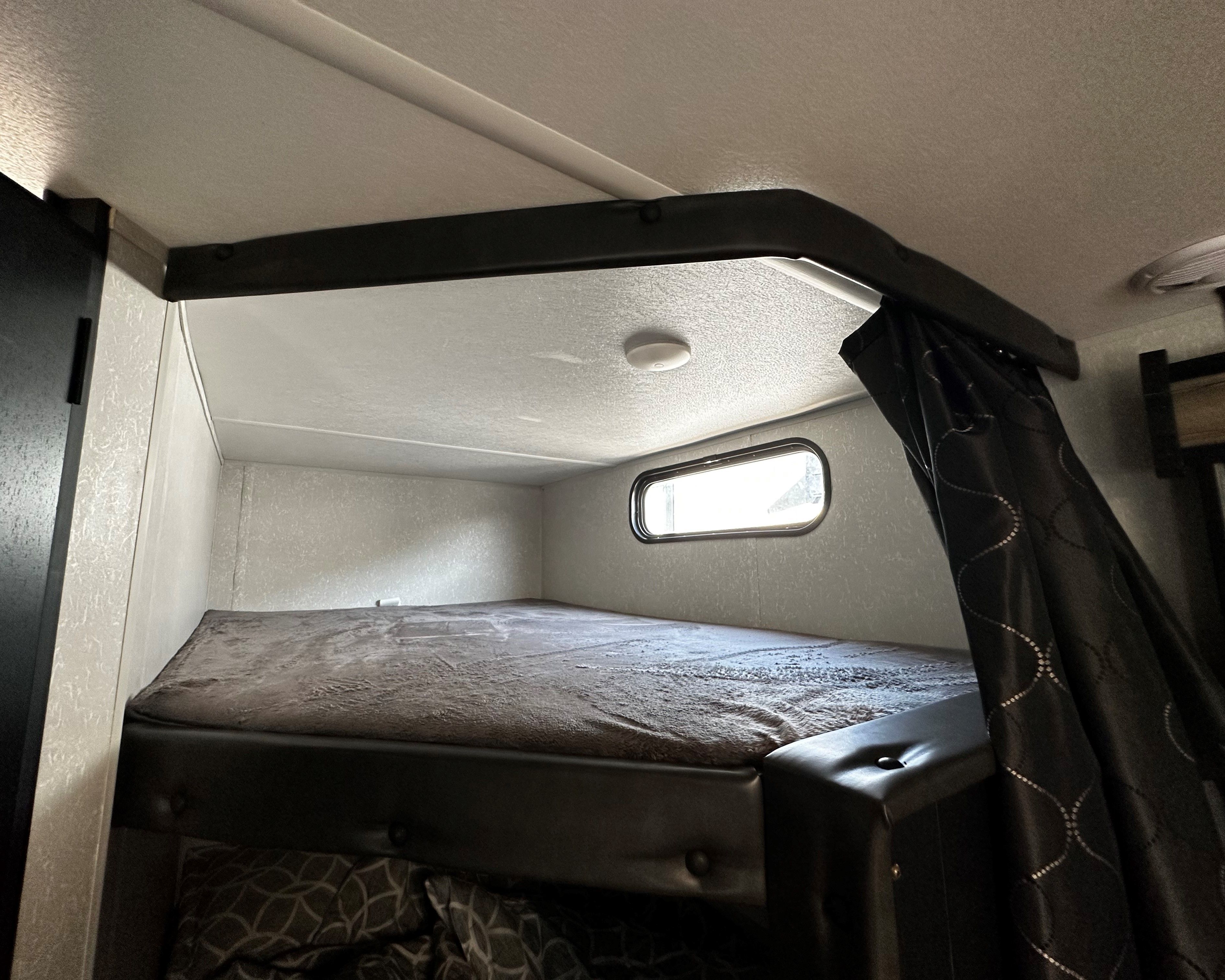 Upper bunk bed in an RV rental, with textured walls, a small window, and a ceiling light, providing a compact sleeping space.. Forest River Cherokee 2021