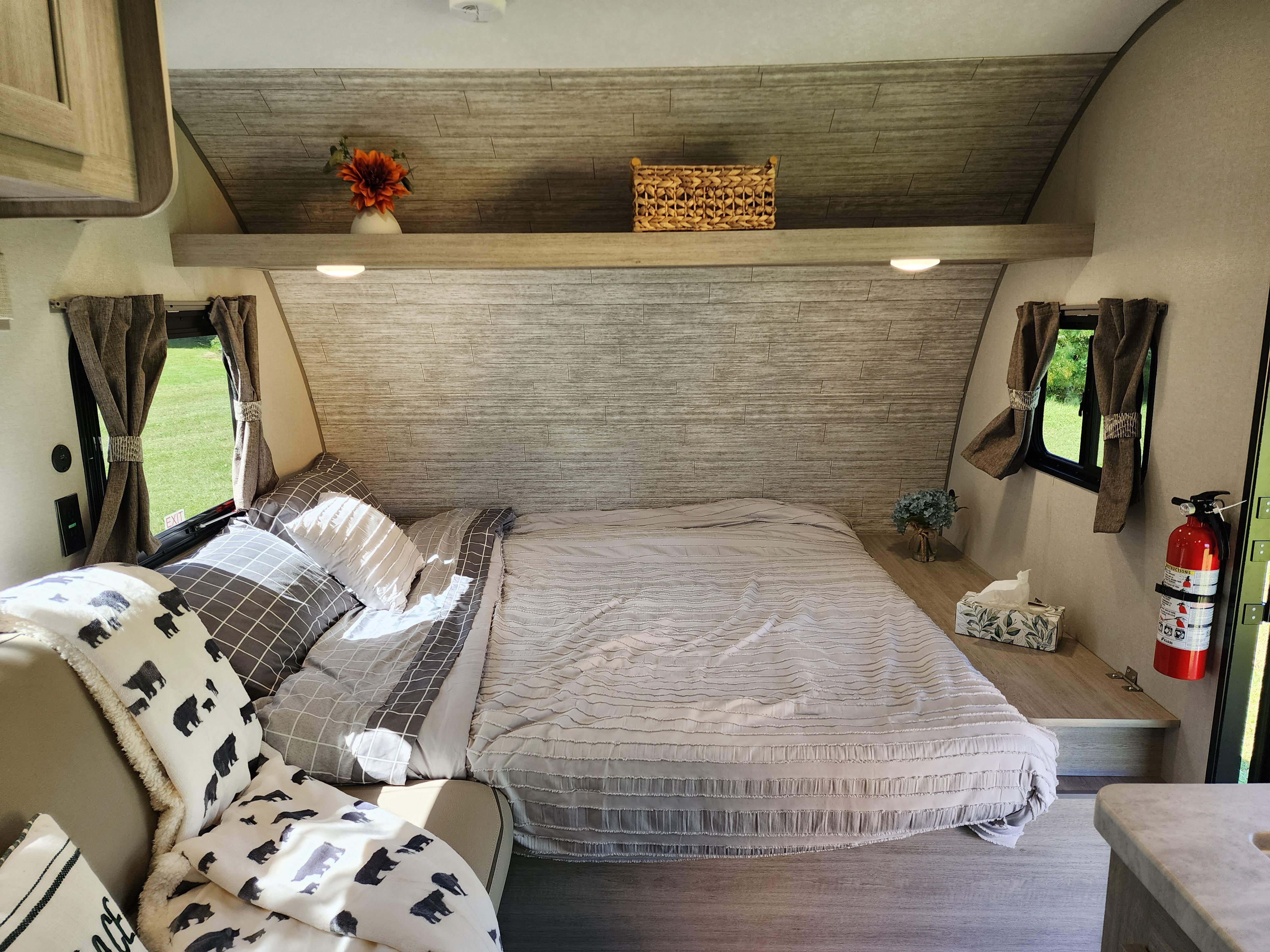 View of a modern RV rental bedroom with a gray bed, bear blanket, wood-look walls, and windows showing green outdoors.. Coachman Catalina Summit Series 7 2024