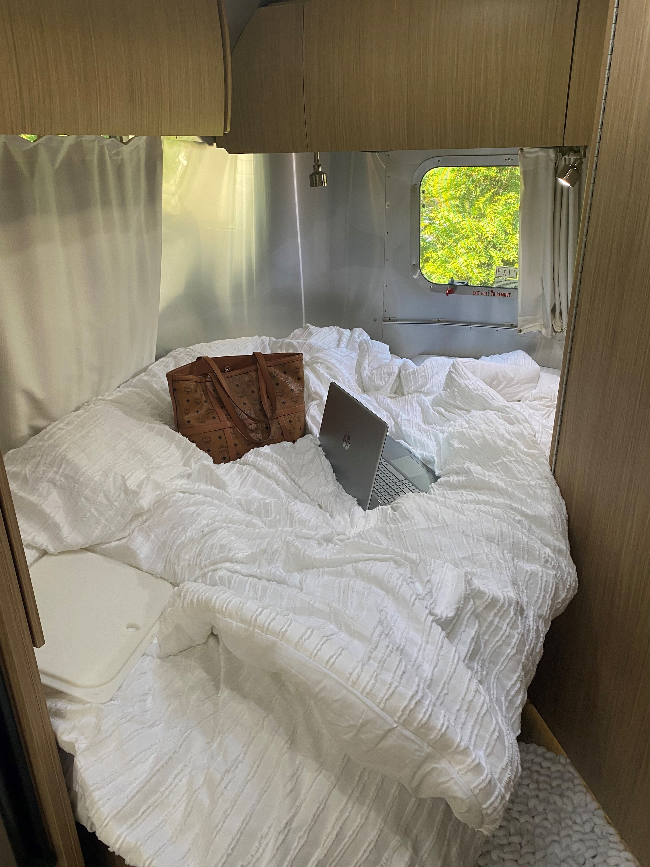 This inviting rv rental features a cozy bed with white duvet, laptop, and an MCM bag, bathed in natural light.. Airstream Flying Cloud 2017