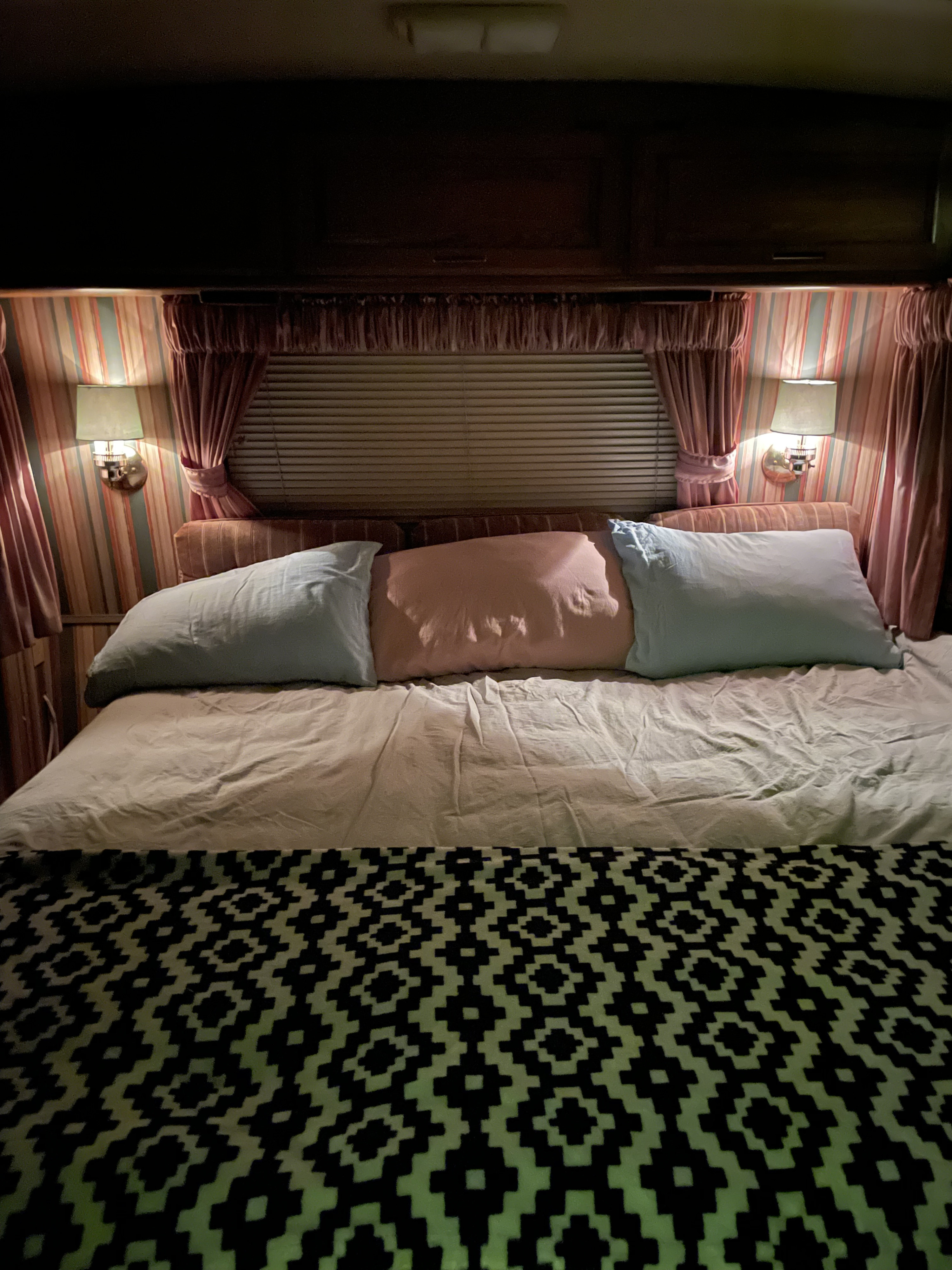 Cozy RV rental bedroom with a bed, three pillows, a geometric patterned blanket, and warm wall sconce lighting.. Winnebago Chieftain 1986