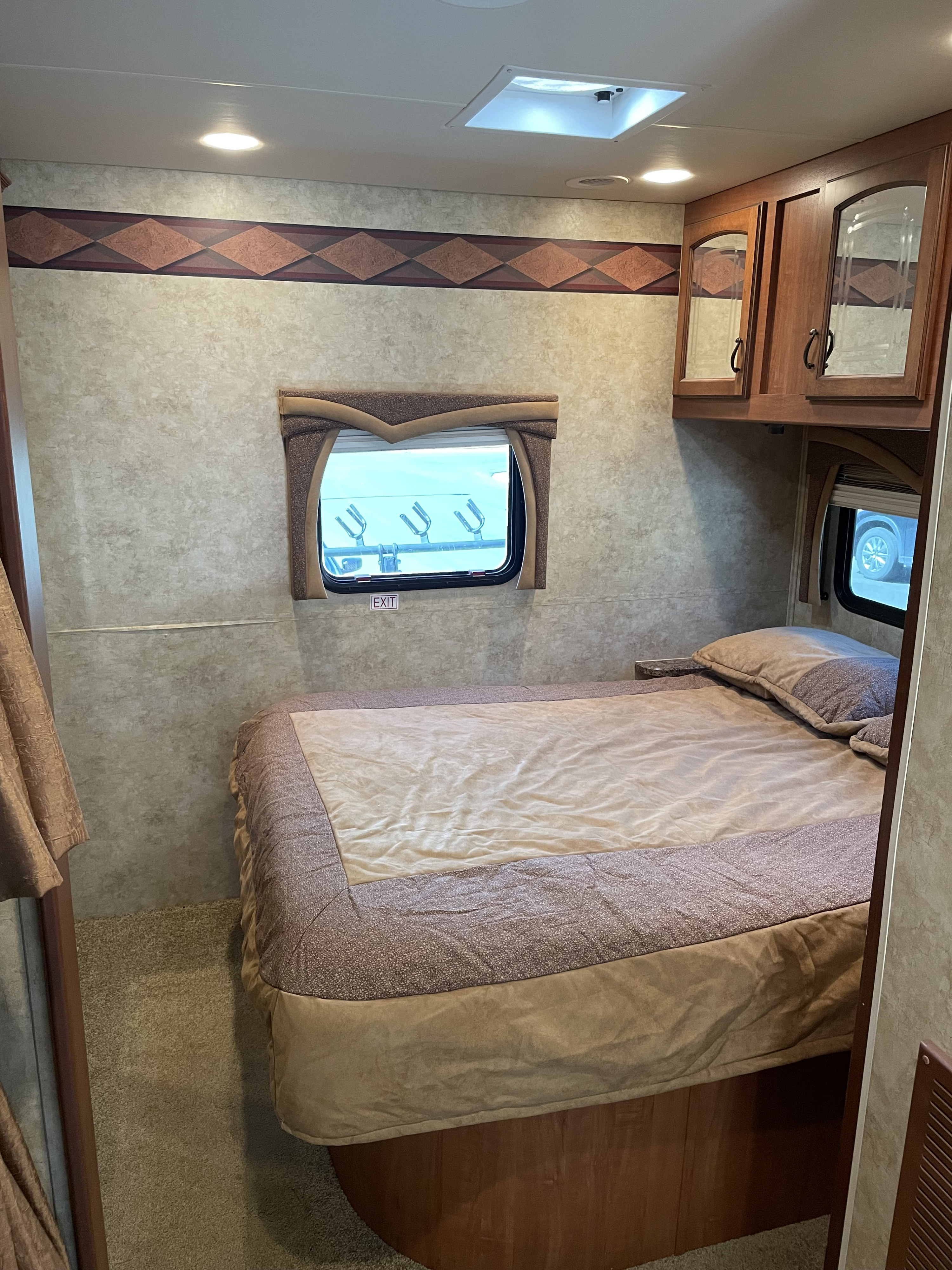 Cozy RV rental bedroom featuring a comfortable bed with light bedding, overhead cabinets, and a window.. Jayco Greyhawk 2010