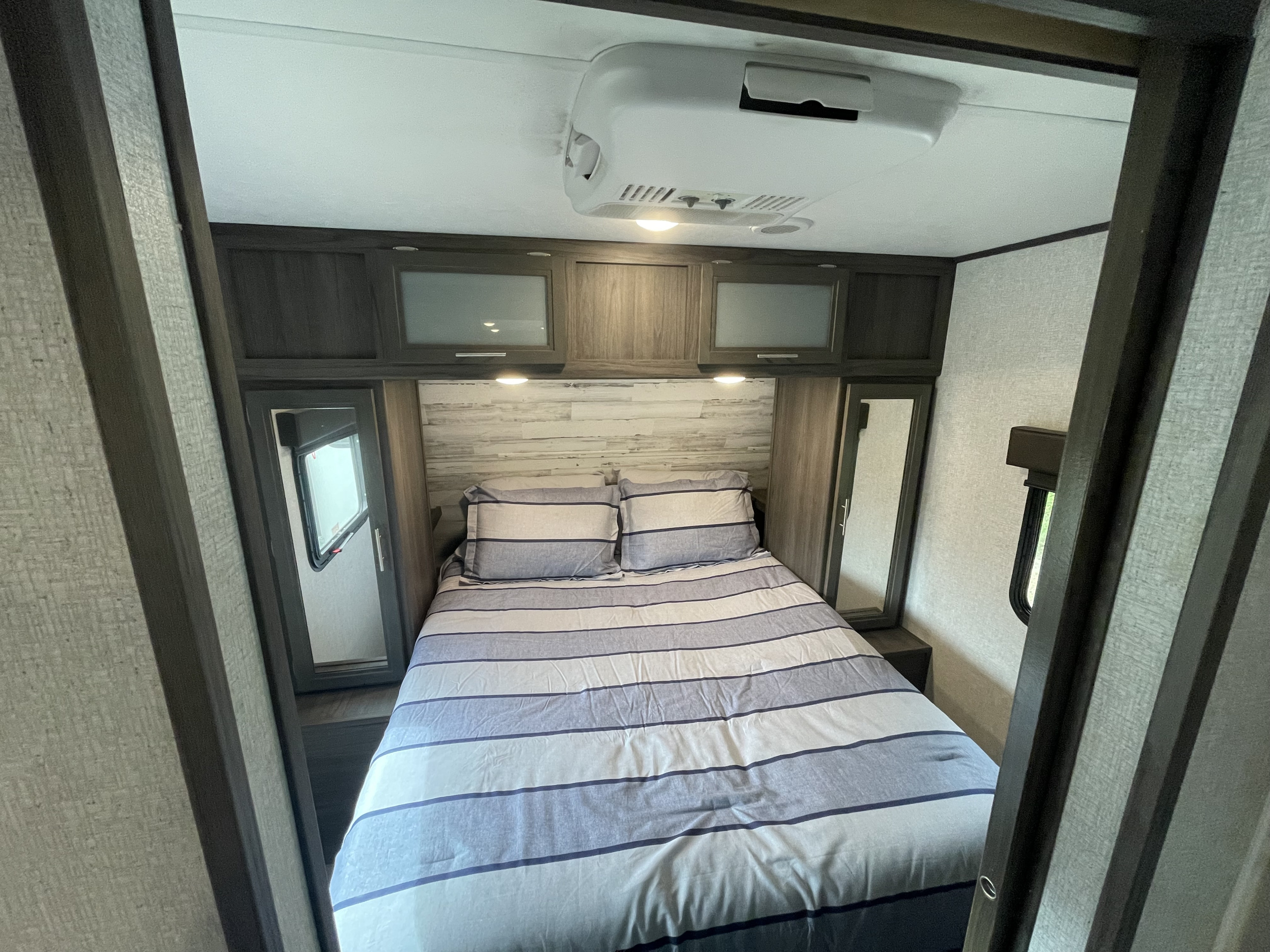 Inside an RV rental: a cozy bedroom with striped bedding, mirrored closets, overhead cabinets, and AC.. Dutchmen Kodiak Ultra Lite 2021