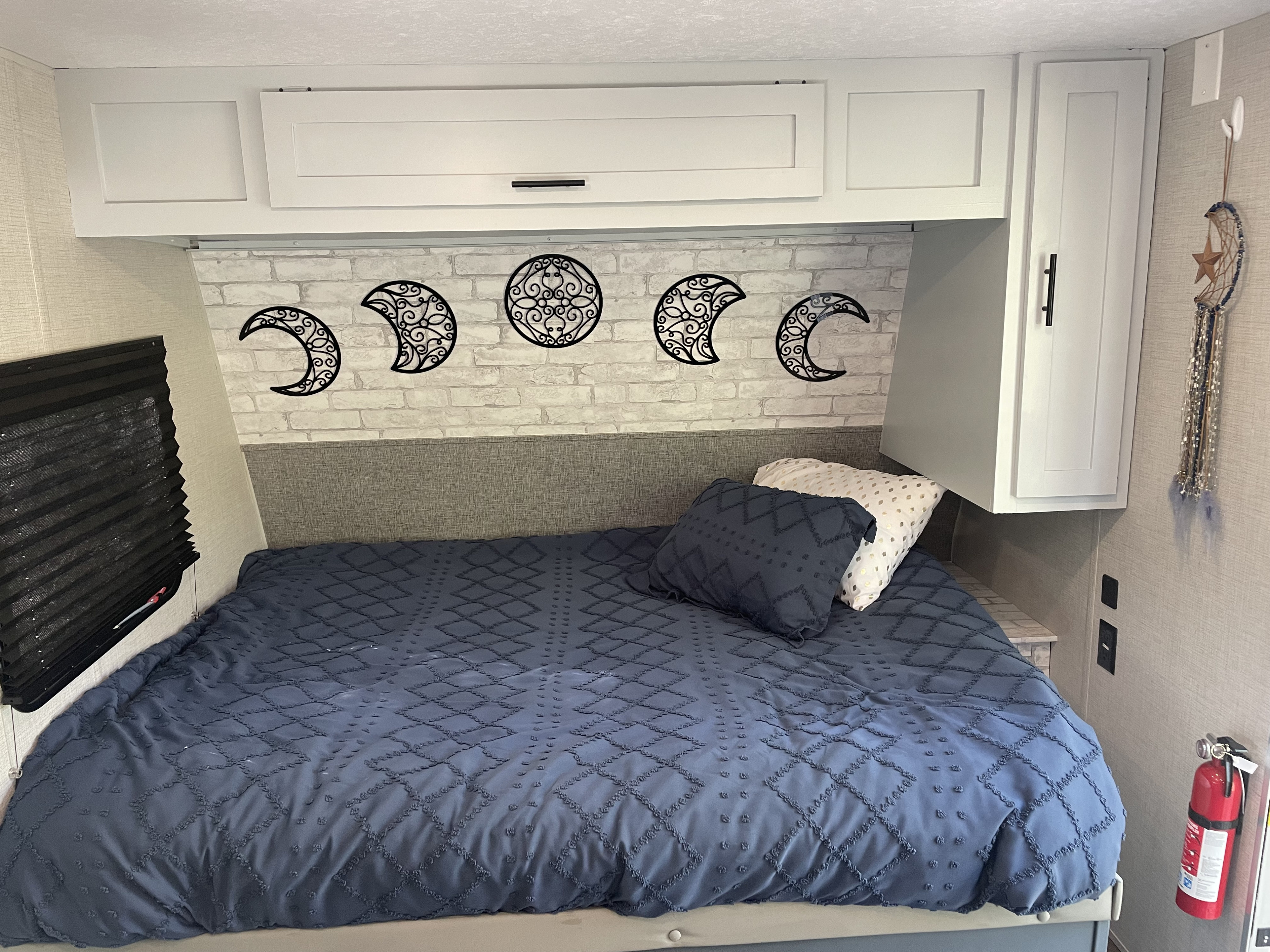 RV rental bedroom: bed with blue comforter, decorative moon phases on brick wall, white cabinets, and fire extinguisher.. Forest River Ozark 1650BHK 2021