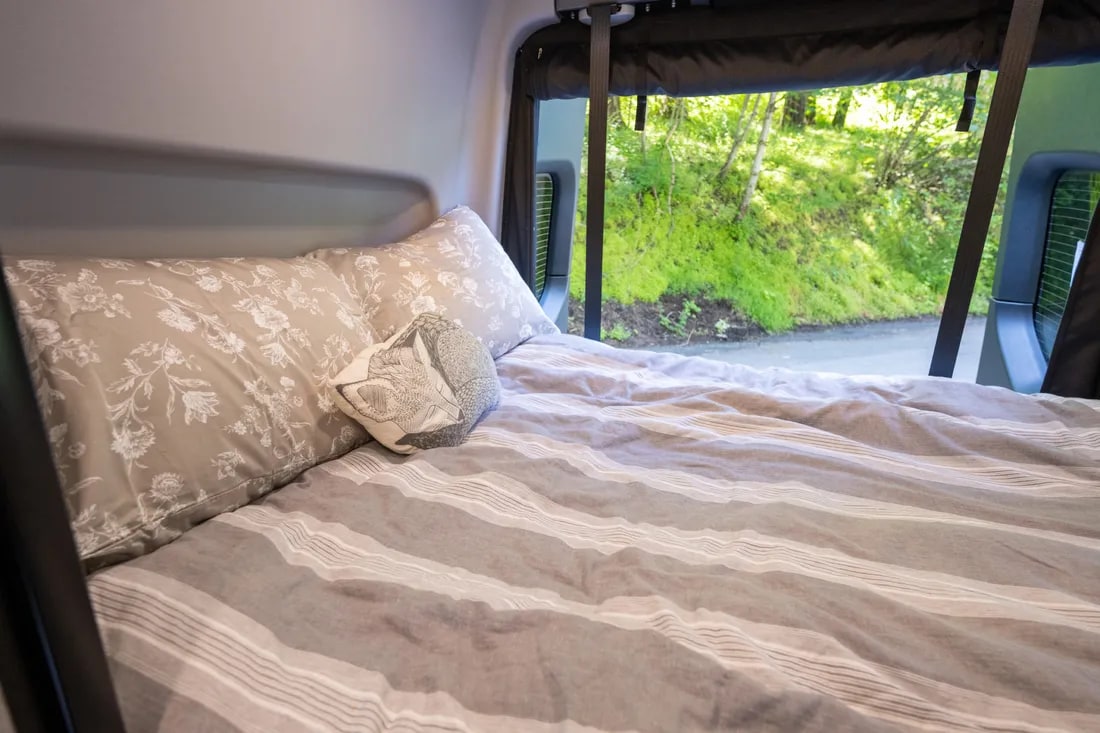 Inside a comfy **RV rental**: a bed with floral pillows, striped duvet, and a scenic window view of nature.. Winnebago Revel 2022