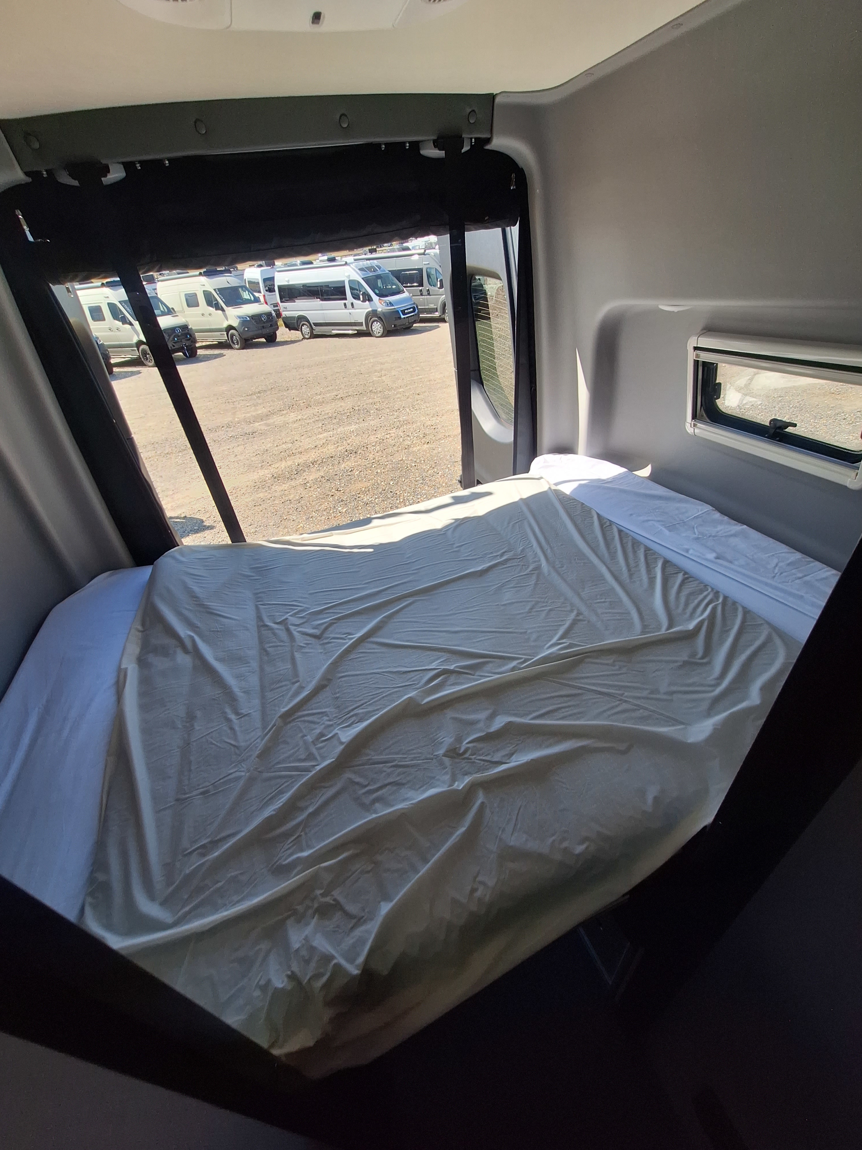 This RV rental's cozy interior features a bed with light sheets, looking out to a lot of parked camper vans.. Winnebago Revel 2023