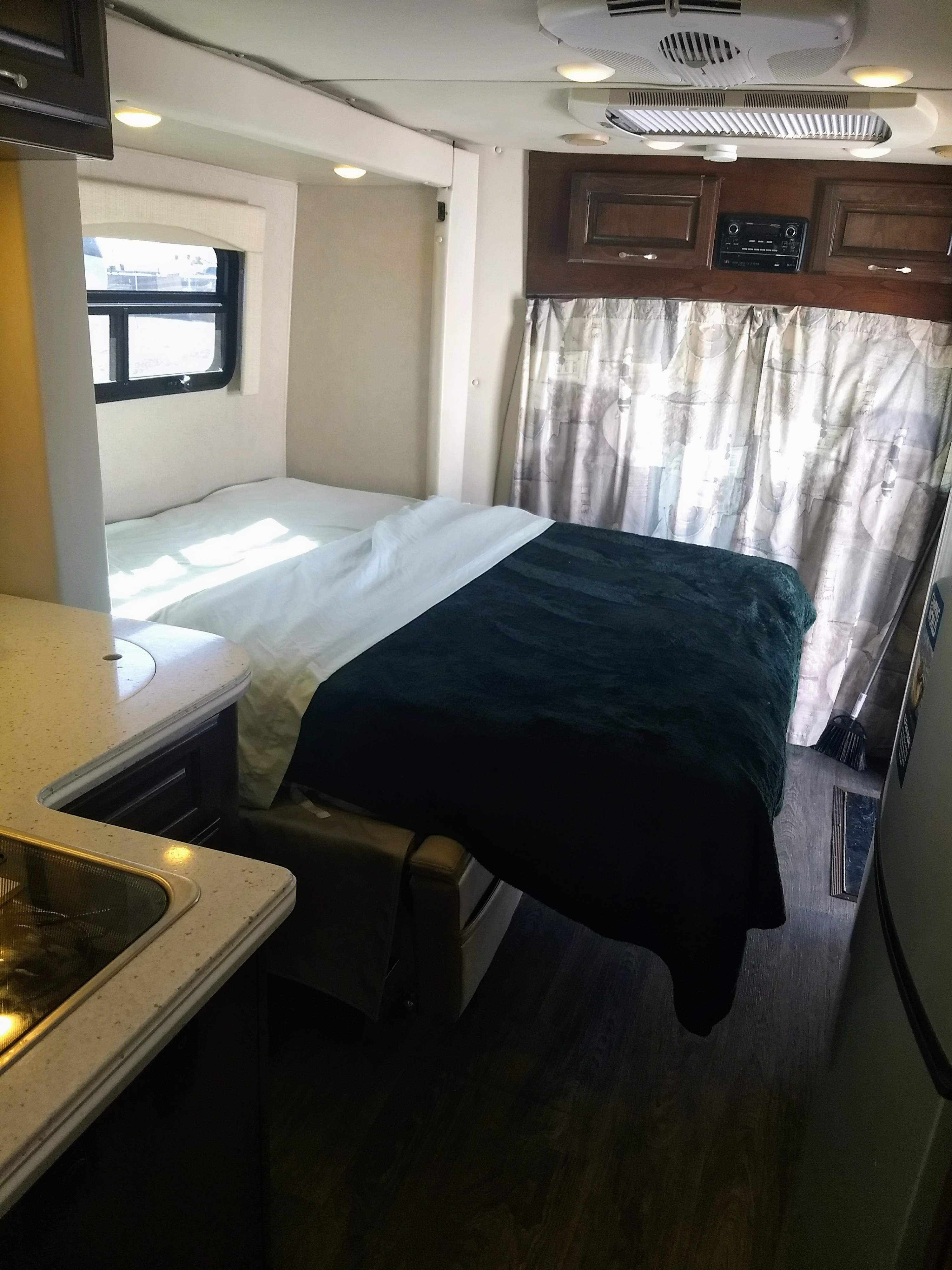 Cozy rv rental interior: A bed with dark green blanket, window, overhead cabinets, and a compact kitchen counter on the left.. Regency Conversions Ultra Brougham 2019
