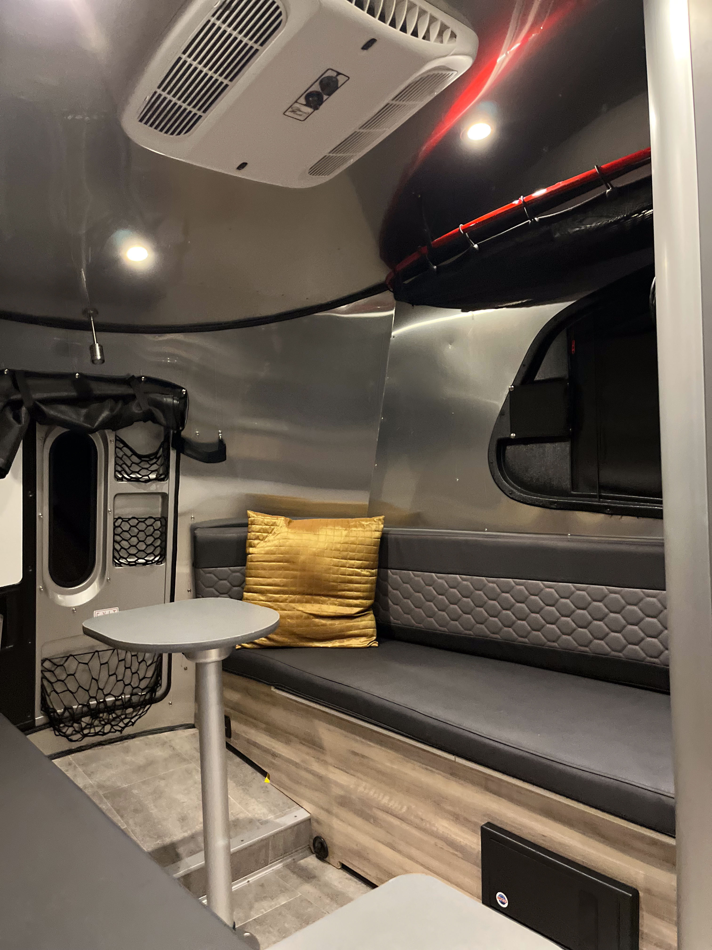 RV rental interior: A modern living space with curved aluminum walls, a grey bench seat, golden pillow, and overhead AC unit.. Airstream Basecamp 2017