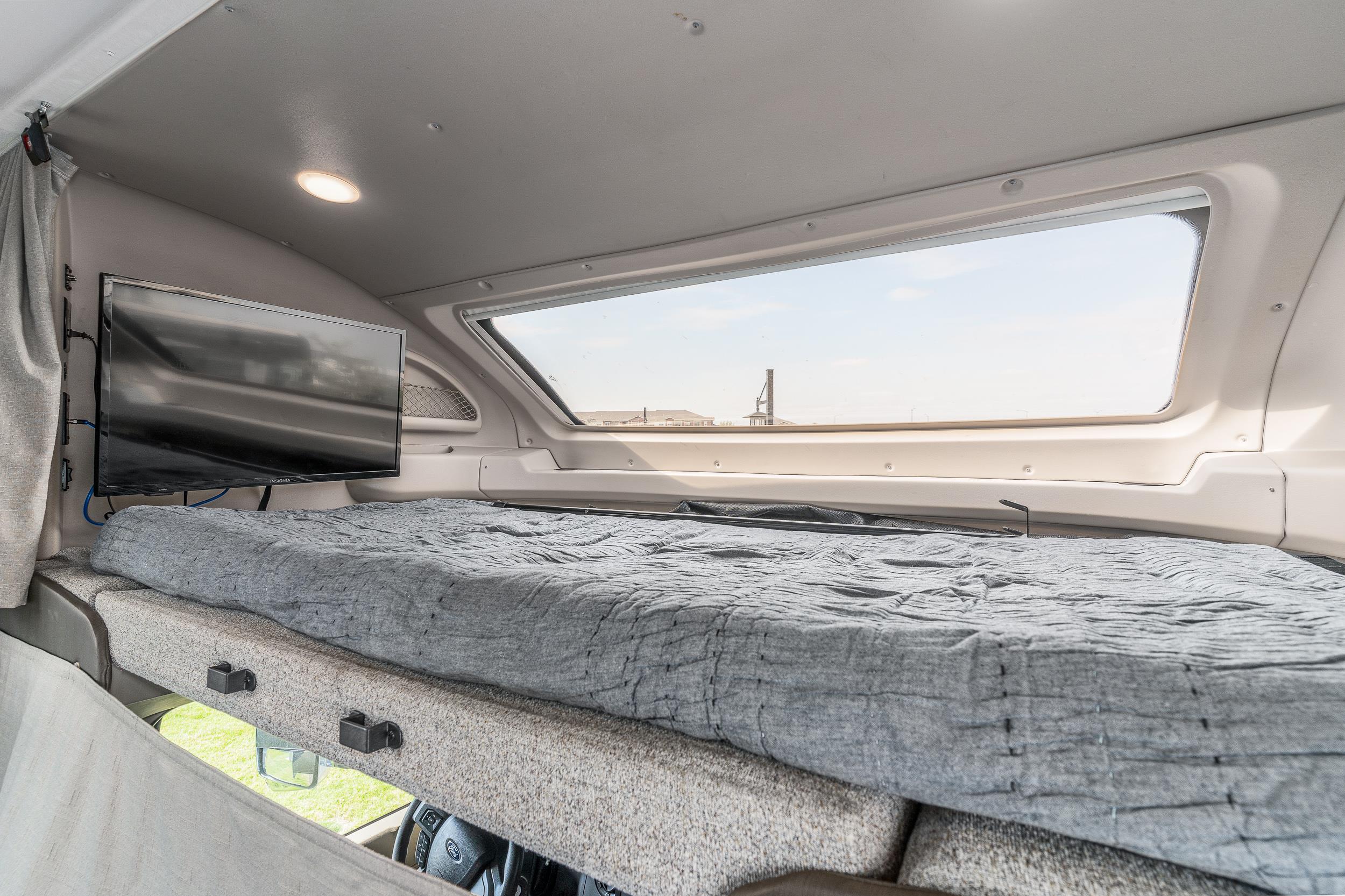 Inside an RV rental: a comfortable overhead bunk bed, flat-screen TV, and large window view.. Jayco Greyhawk Prestige 2022