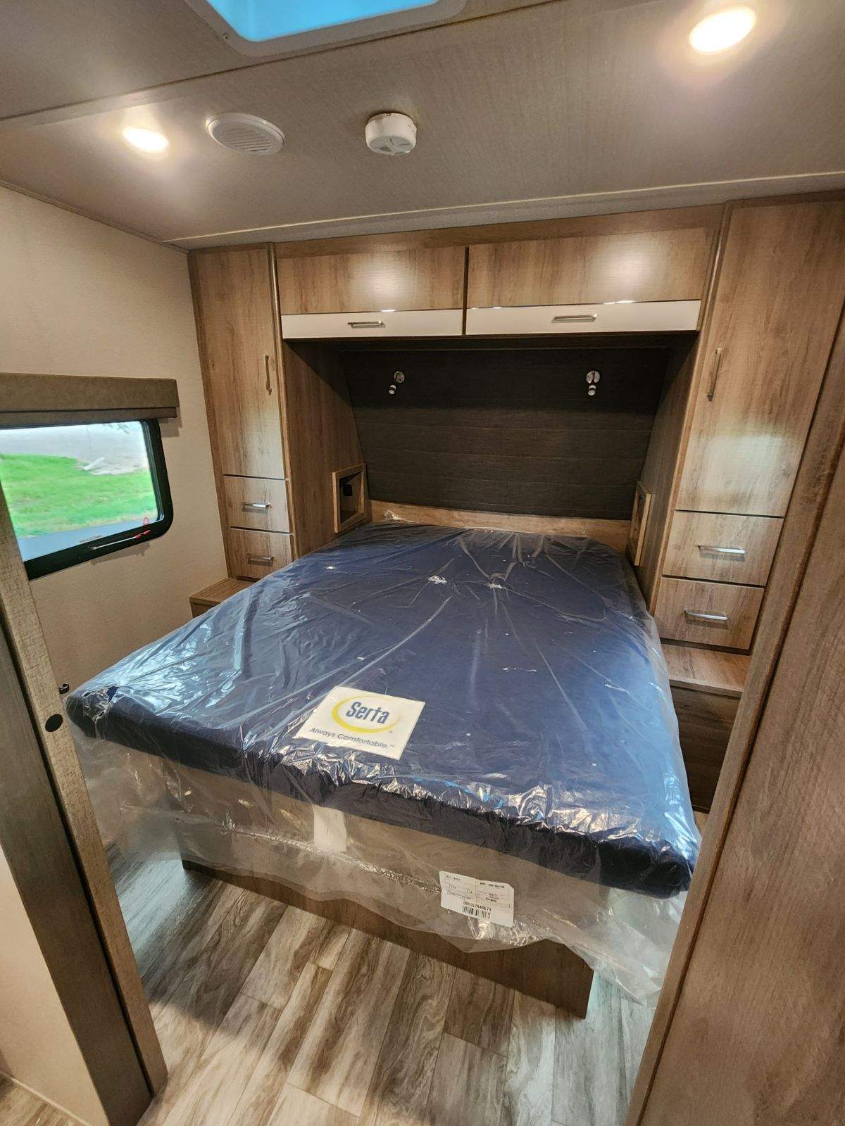 Compact RV rental bedroom featuring a Serta bed (in plastic), built-in storage, and a window.. Grand Design Imagine 2022