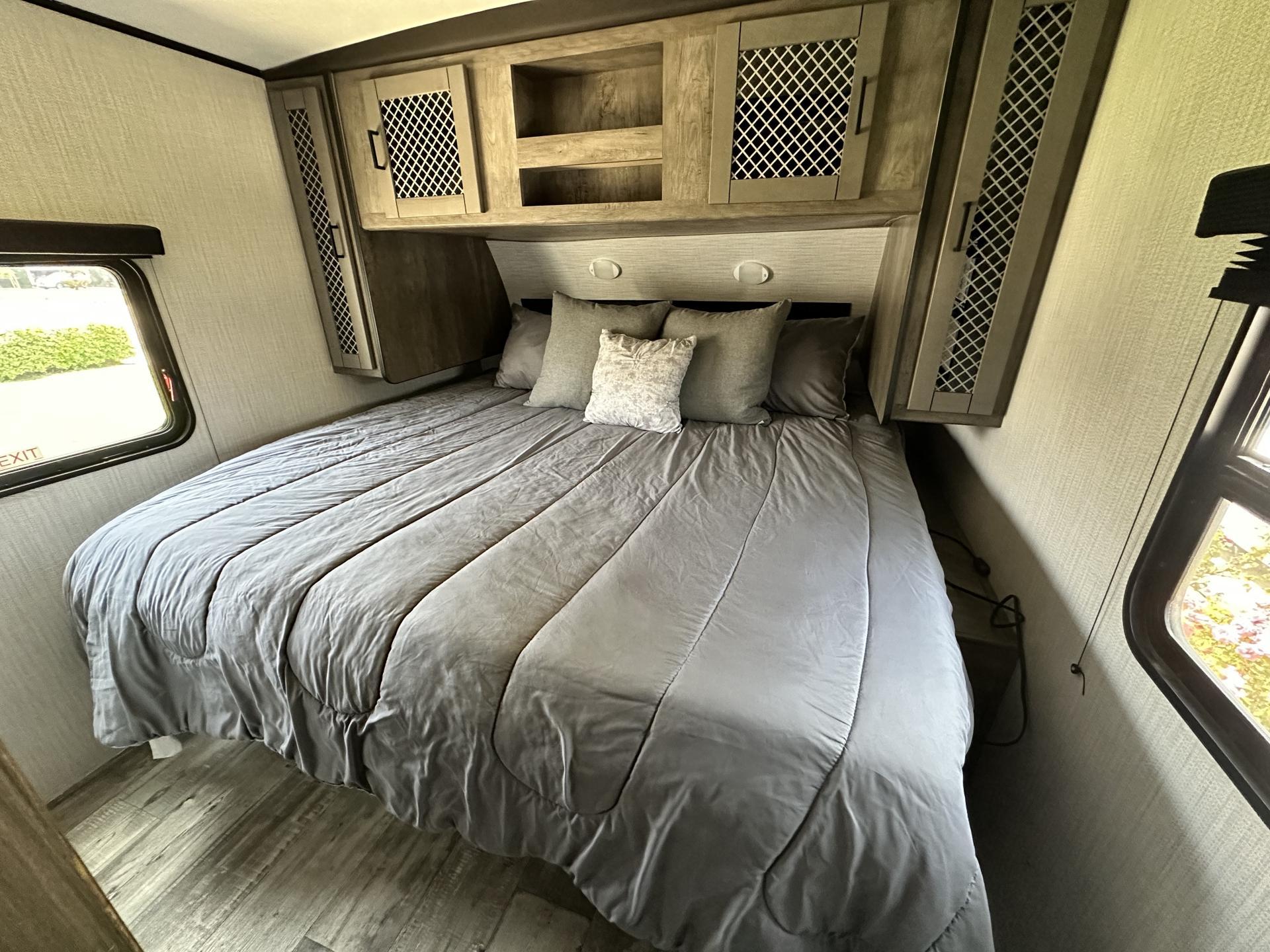 Interior of an RV rental bedroom showing a gray-comforter bed, built-in overhead cabinets, and side windows.. Heartland RVs North Trail 2021
