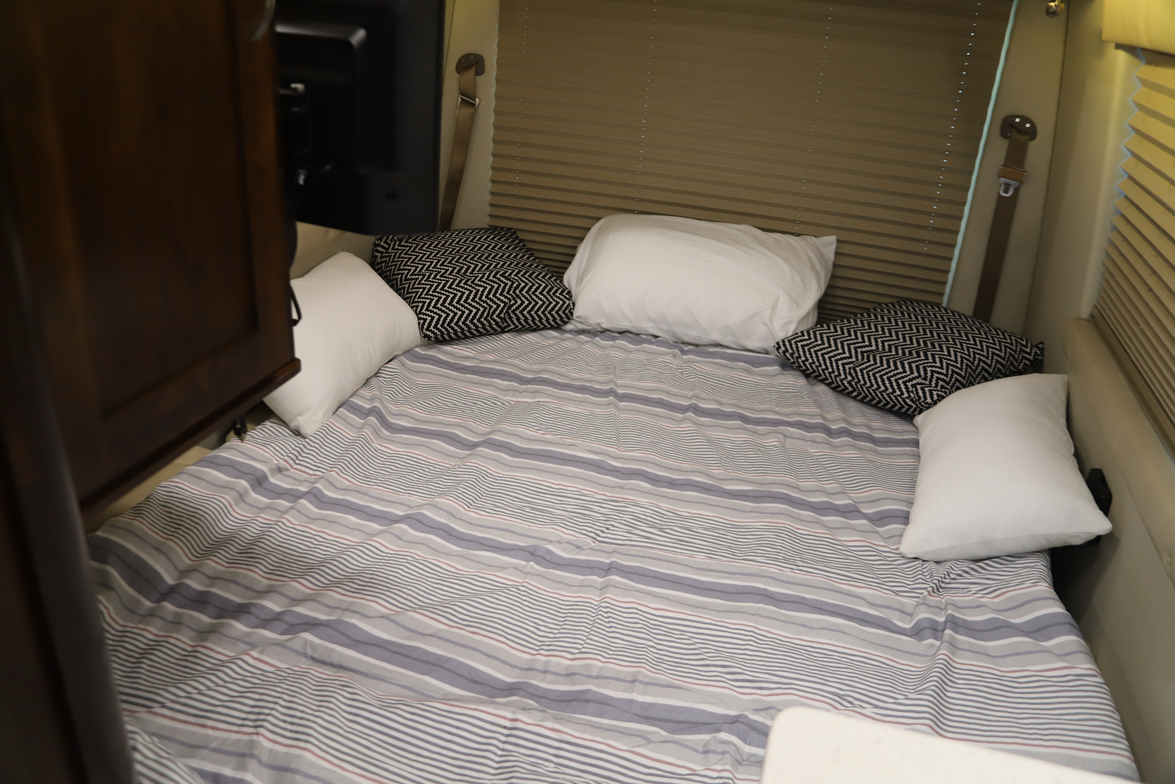 Relax in this cozy RV rental bed featuring striped bedding, white and geometric pillows, and pleated window shades.. Forest River Coachmen Galleria 2019