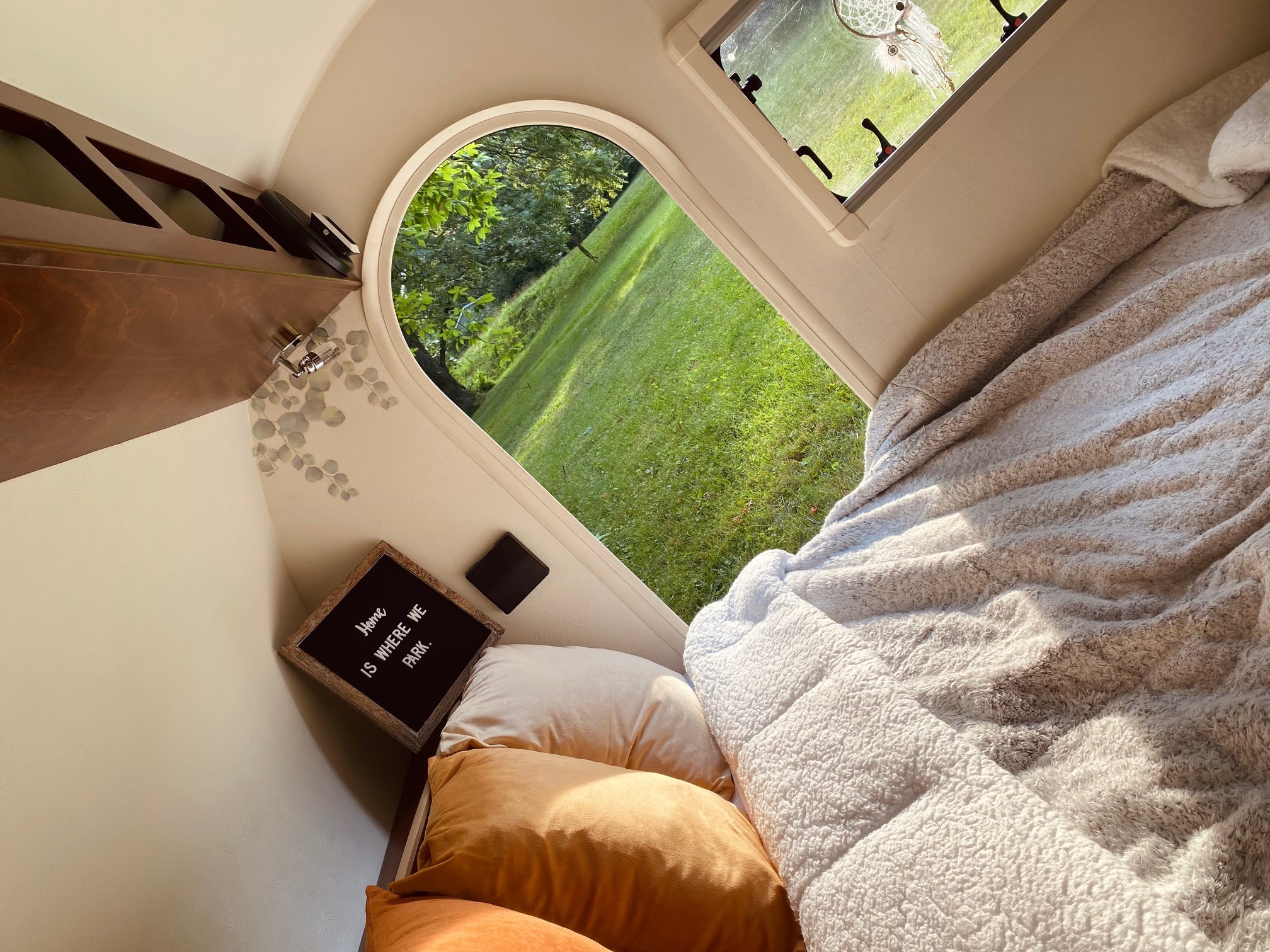 Cozy RV rental interior featuring a comfy bed with a soft blanket, an arched window offering a scenic view of a green field, and a "Home is where we park" sign.. Little Guy T@G Little Guy T@G Trailer 2016