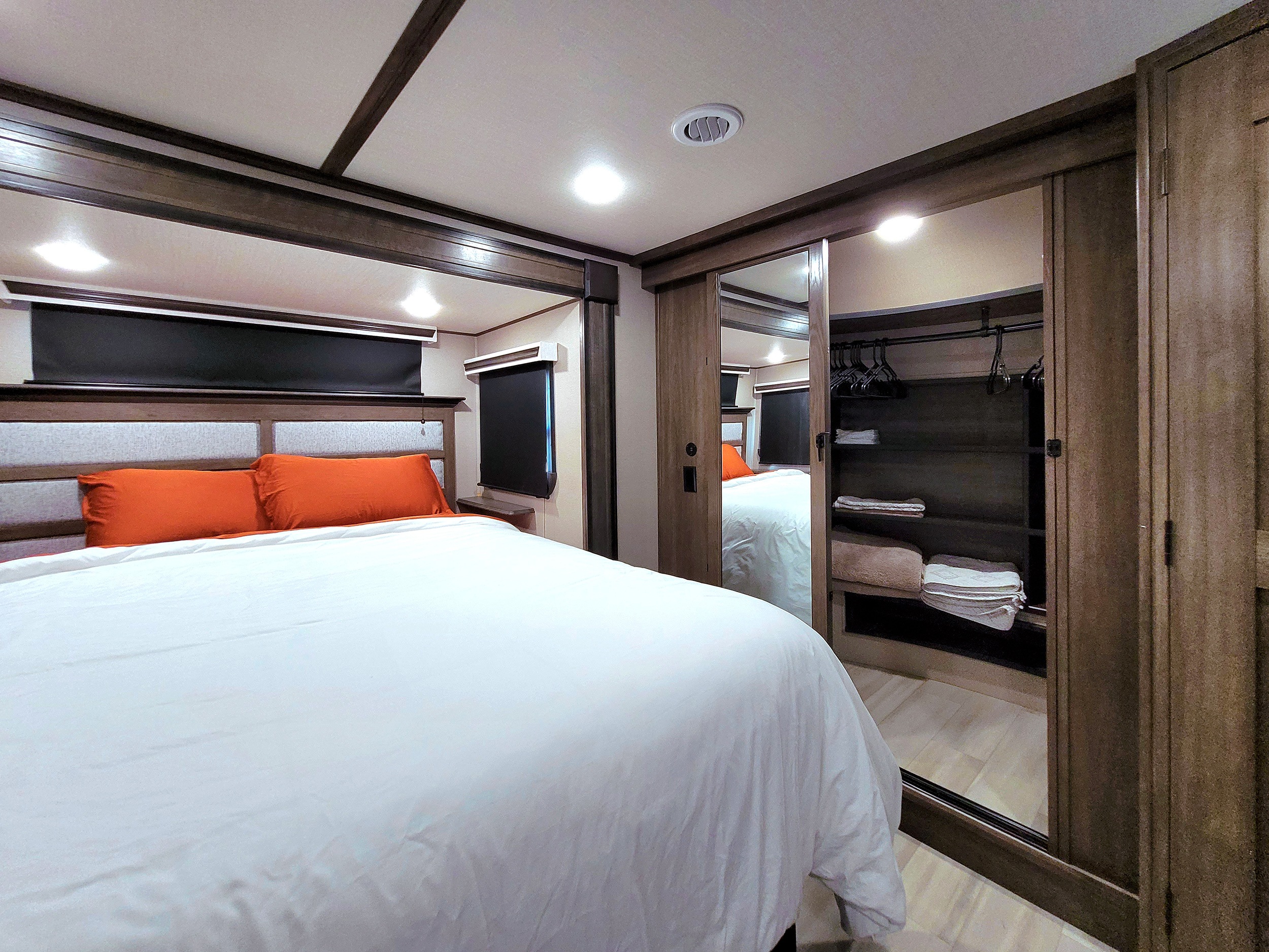 Discover comfort in this RV rental bedroom, featuring a plush bed, orange pillows, window blinds, and mirrored closet with storage.. Grand Design Solitude 2022