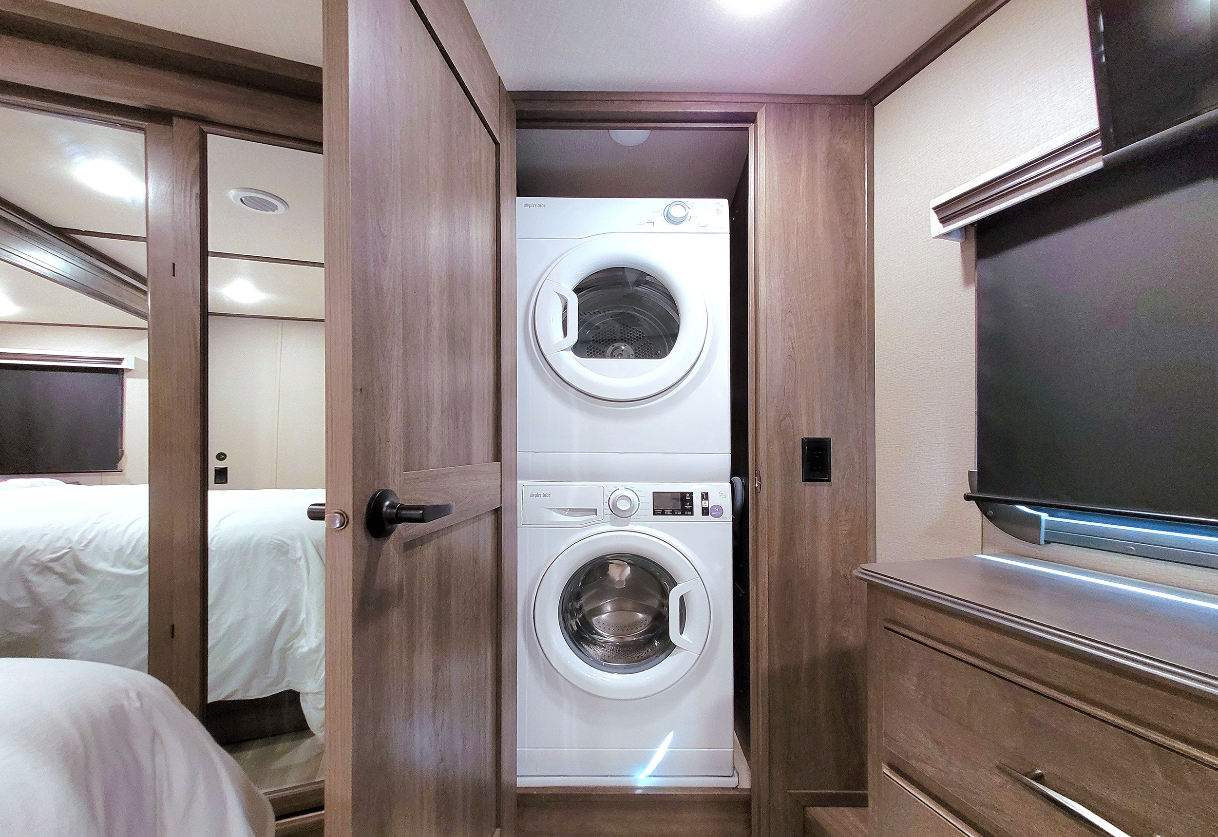 Stacked washer/dryer in an RV rental, featuring a comfortable bedroom with a TV and warm wood finishes.. Grand Design Solitude 2022