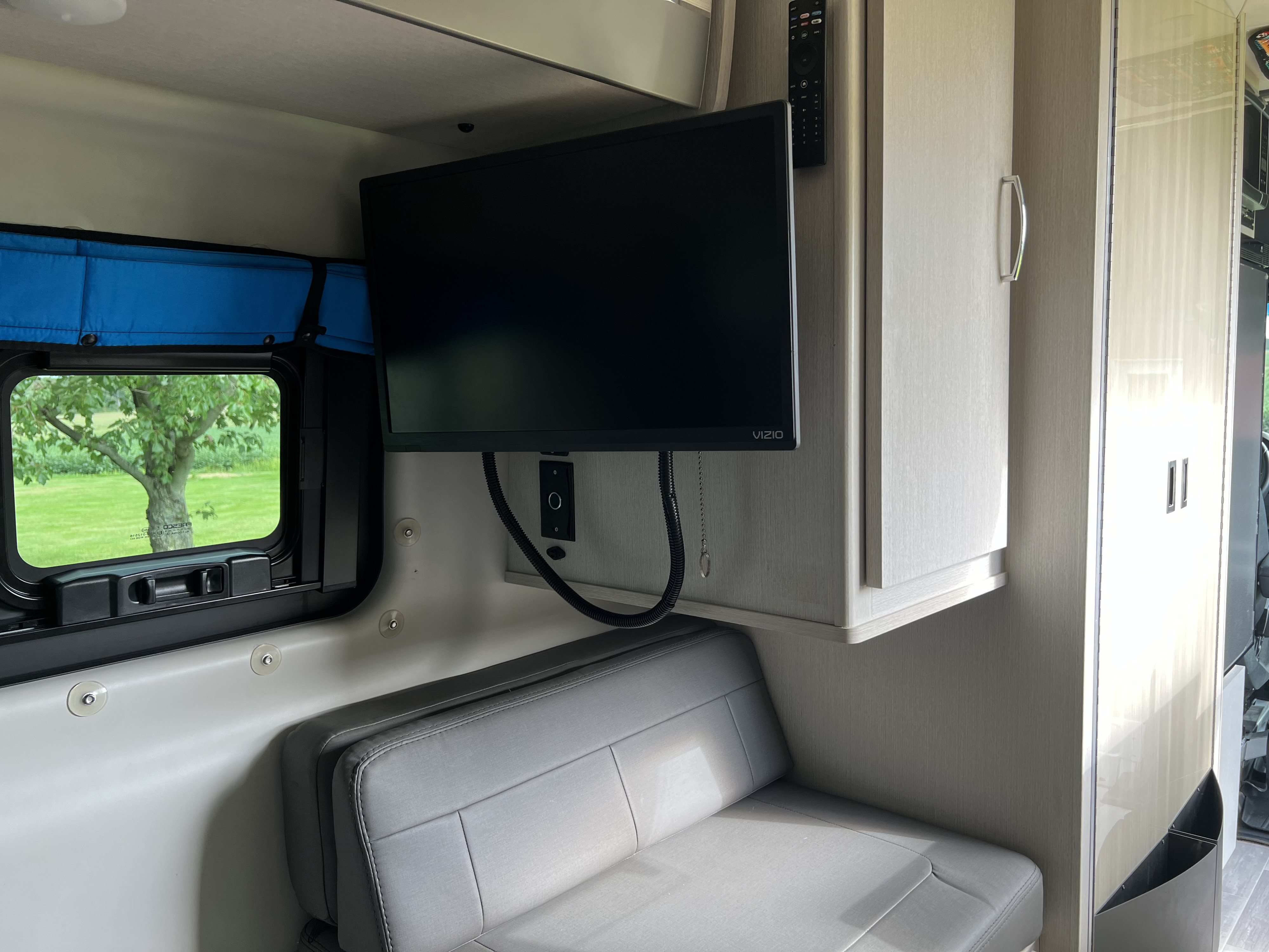 Inside an RV rental: a Vizio TV mounted above a cozy bench seat, with a bright window overlooking green grass.. Thor Sanctuary 2023