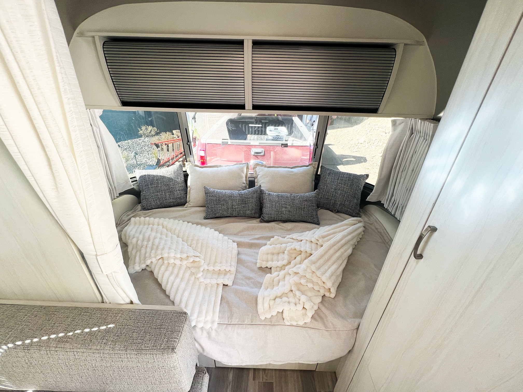 Step into comfort in this RV rental bedroom: a large bed with pillows, soft blankets, and windows overlooking a red truck.. Airstream Bambi 2023