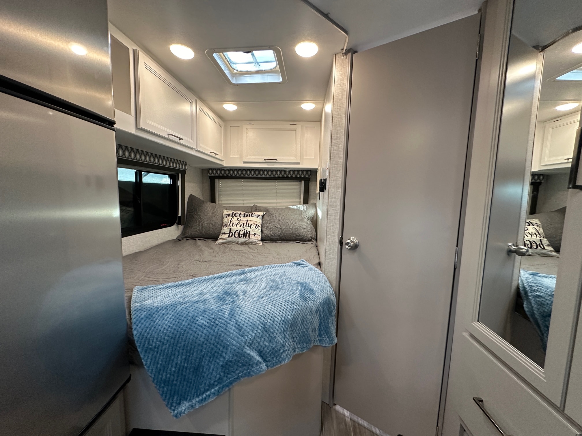 Cozy RV rental bedroom area with a bed, blue blanket, overhead cabinets, and a partial view of a stainless steel fridge.. Thor Geneva 22VT 2024