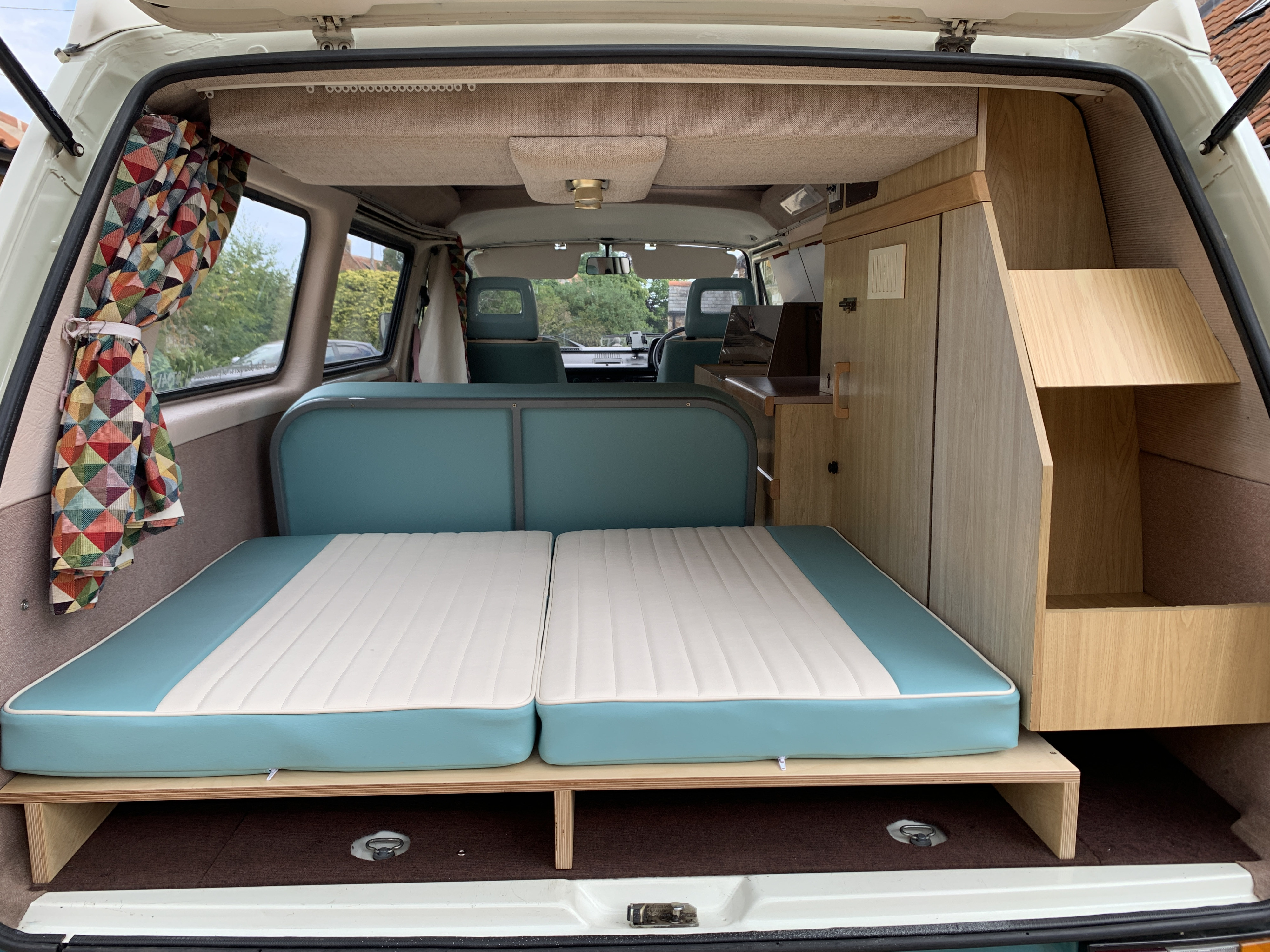 Interior of an RV rental: a cozy rear view with blue and white bed cushions, colorful curtains, and wooden cabinetry.. Volkswagen Campervan 1989