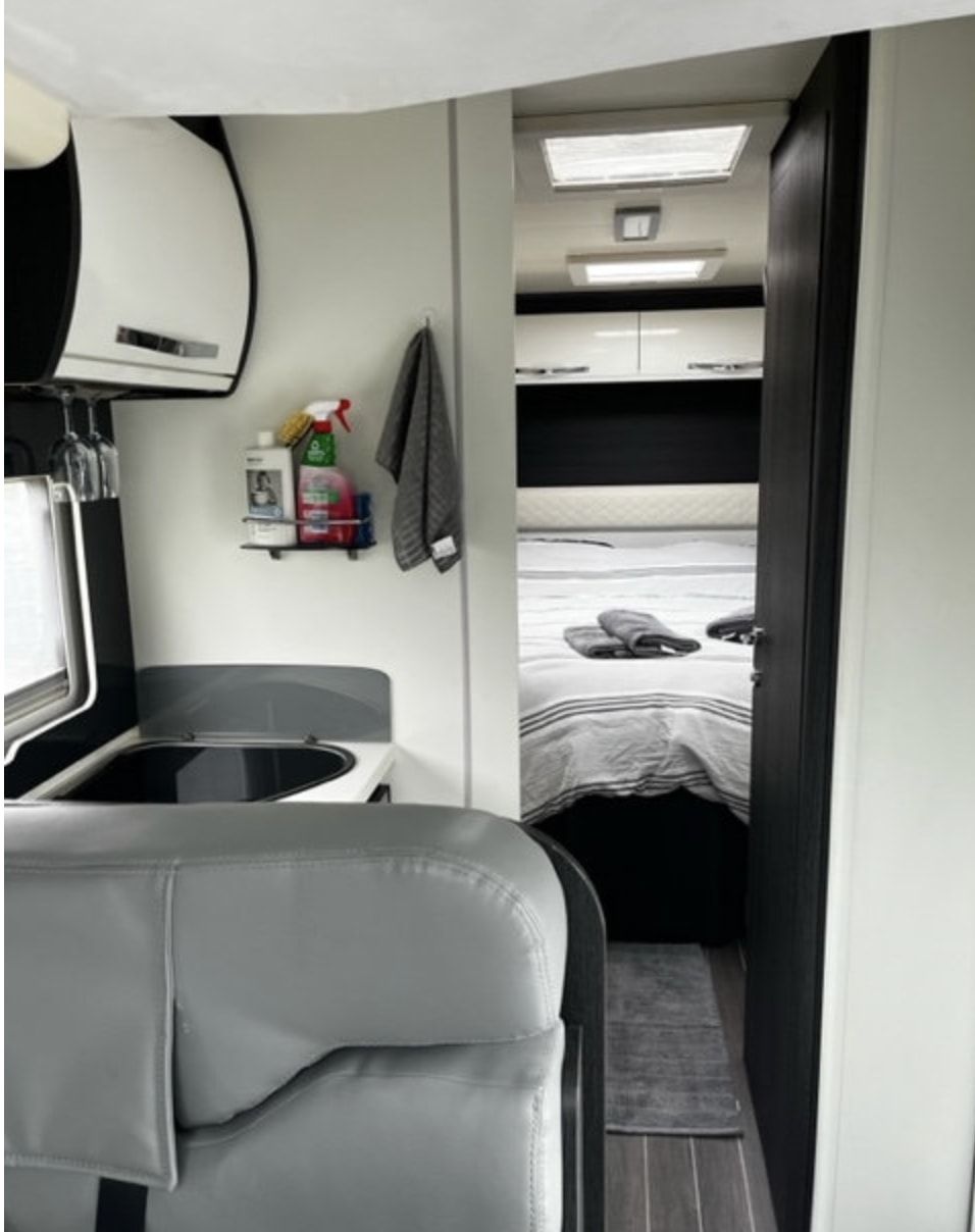 Inside a modern RV rental: a gray upholstered seat in the foreground, leading to a compact kitchen and then a cozy bedroom.. Ford Rollerteam 696 2019