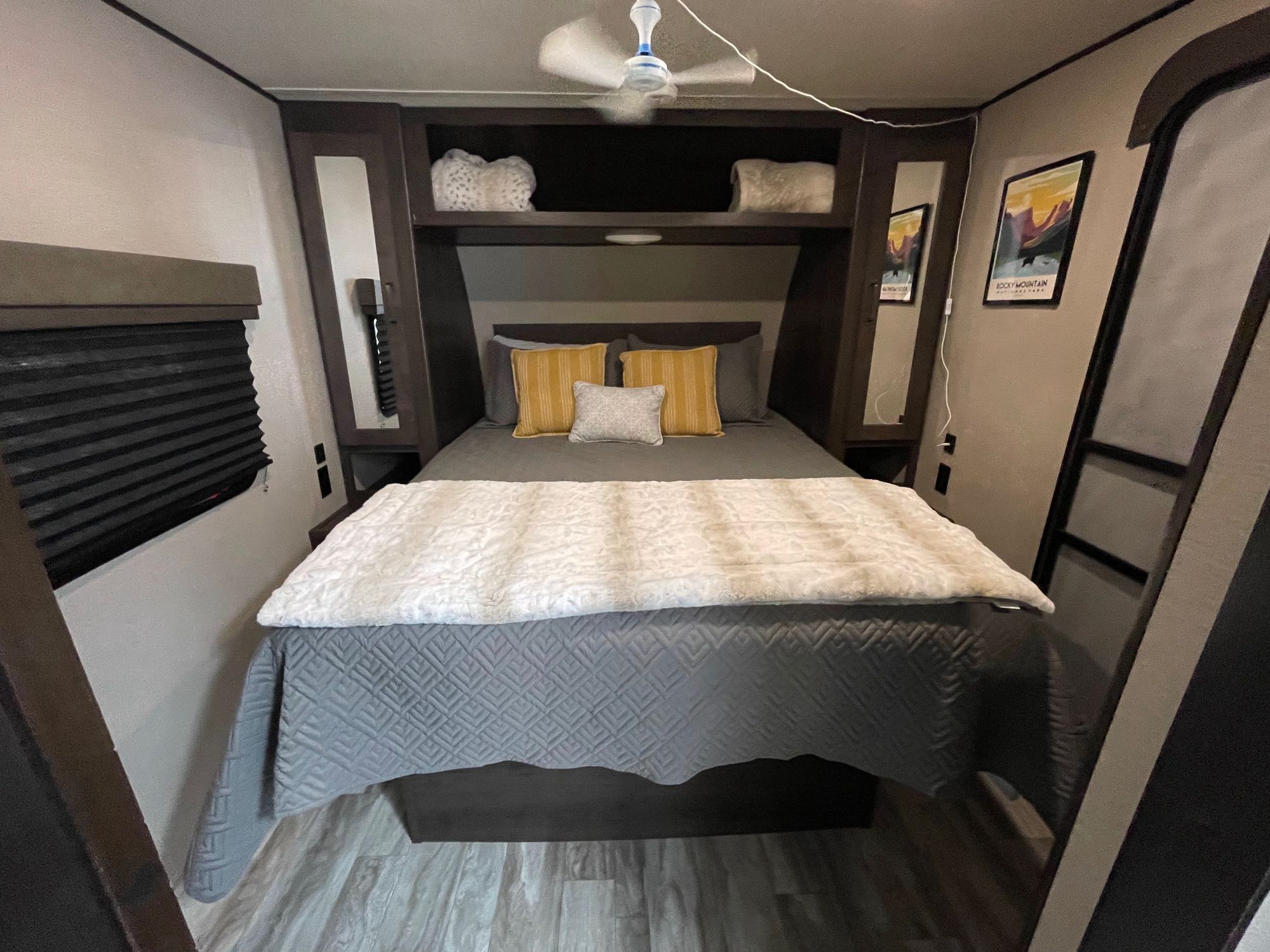 Explore this cozy RV rental bedroom! Featuring a bed with gray bedding, built-in shelves, ceiling fan, and a Rocky Mountain art print.. Grand Design Transcend Xplor 2022