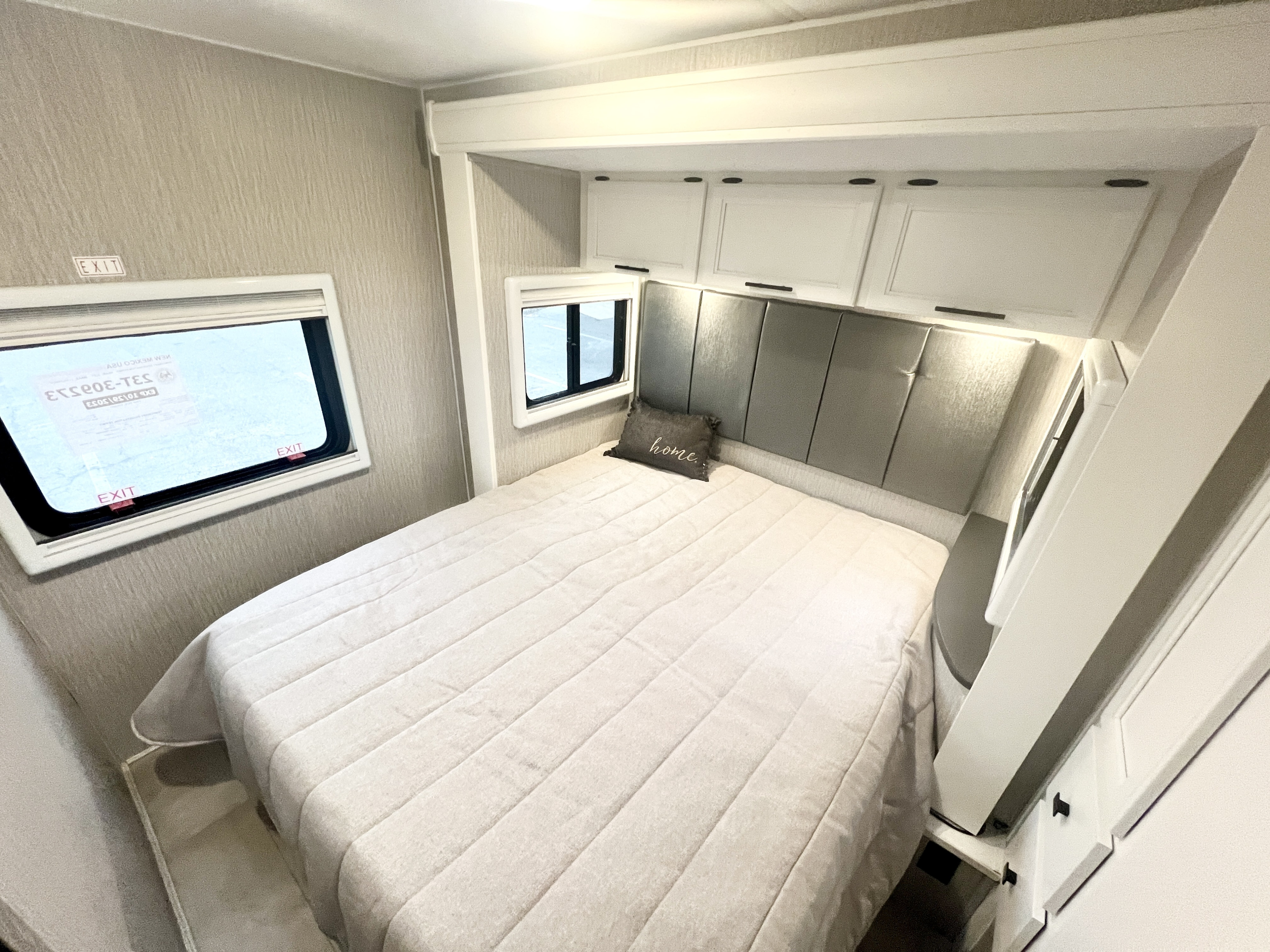 Cozy RV rental bedroom with a soft, light bed, padded gray headboard, white overhead cabinets, and windows.. Coachmen Prism 2024