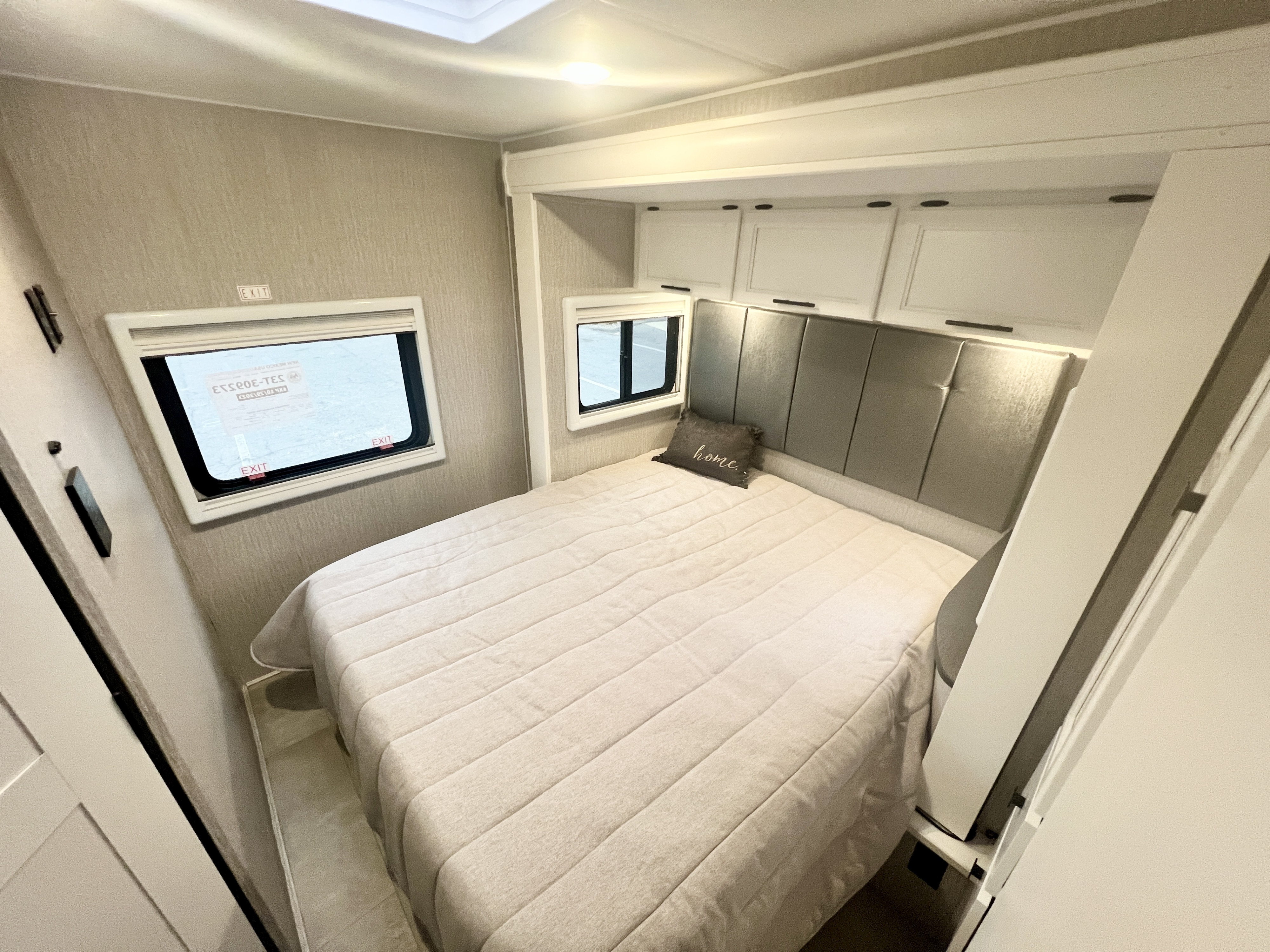 This rv rental shows a compact bedroom with a bed, padded headboard, overhead cabinets, and windows on textured walls.. Coachmen Prism 2024