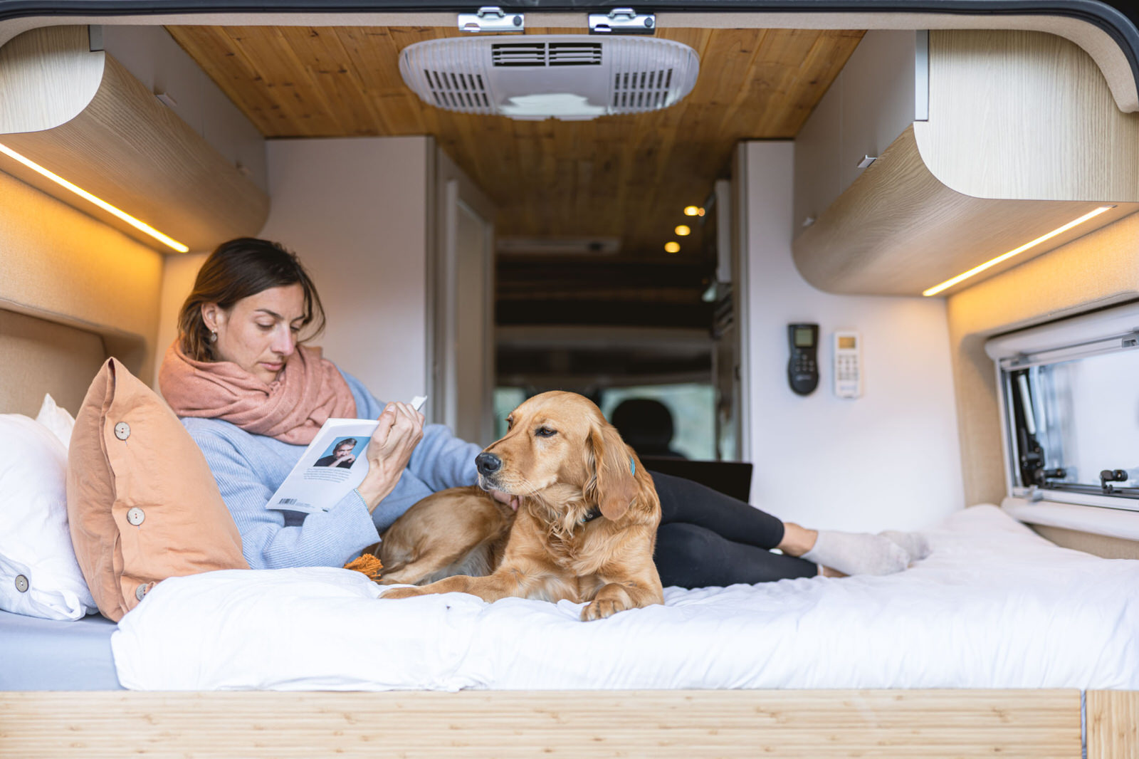 Cozy RV rental life: Woman reading a book beside her golden retriever on a bed inside a modern camper.. Noovo Lite 2023