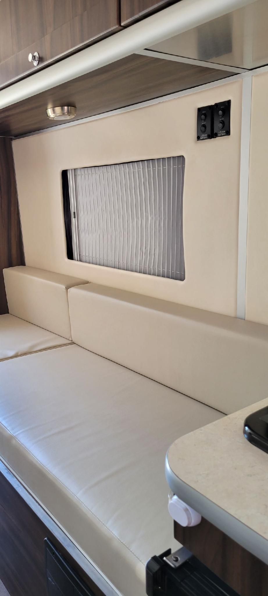 Light-colored upholstered bench seating and window with pleated blind inside an RV rental. Overhead cabinets and battery controls visible.. Carado Carado Class B 2018