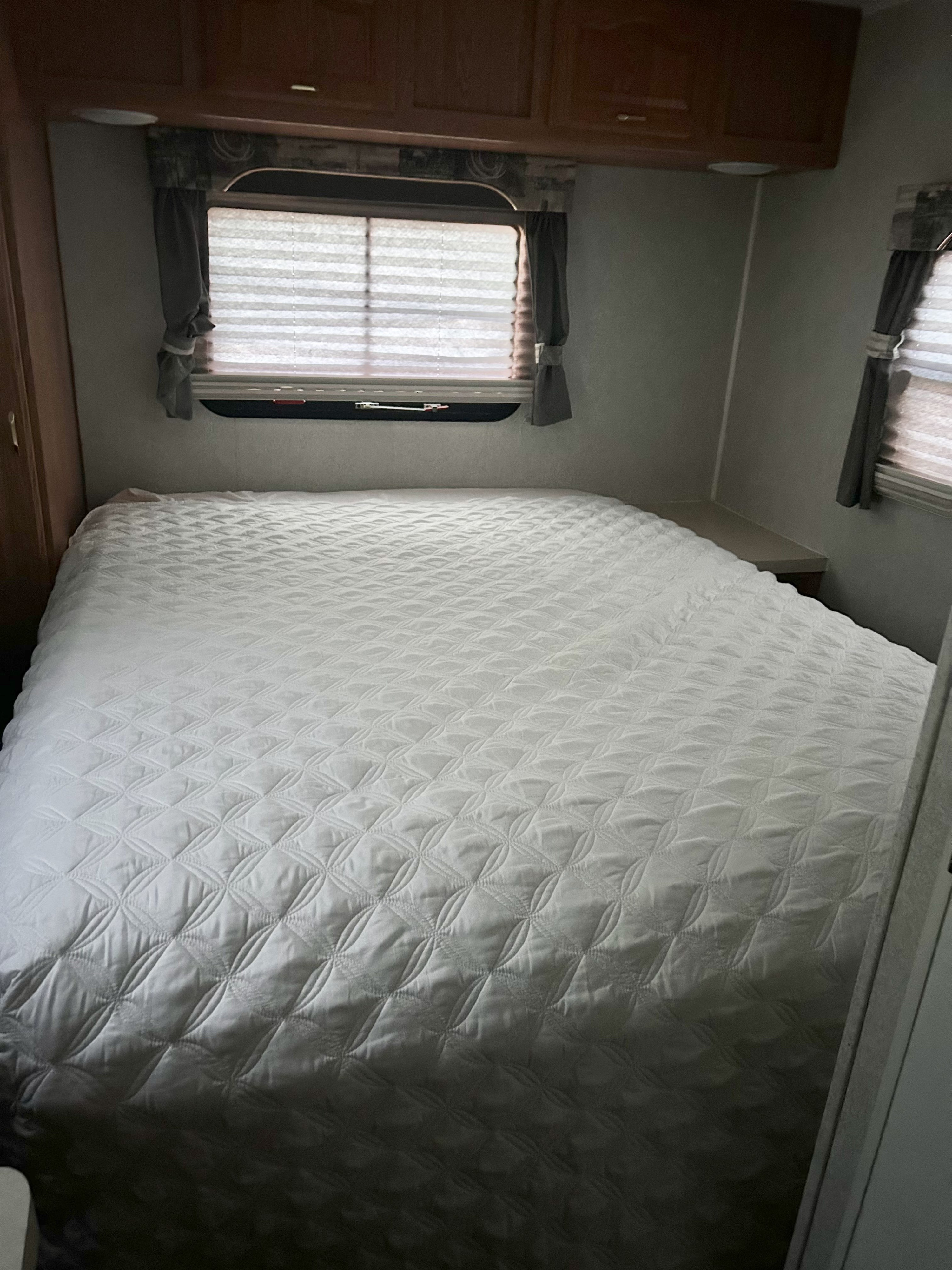 Compact RV rental bedroom featuring a white quilted bed, overhead storage, and a window with blinds and curtains.. Forest River Rockwood Ultra Lite 2003
