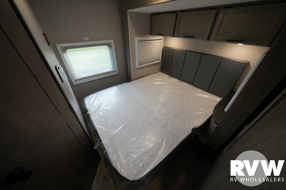 Inside an RV rental: a cozy bedroom with a plastic-wrapped mattress, overhead cabinets, and a window for natural light.. Coachmen Prism 2024