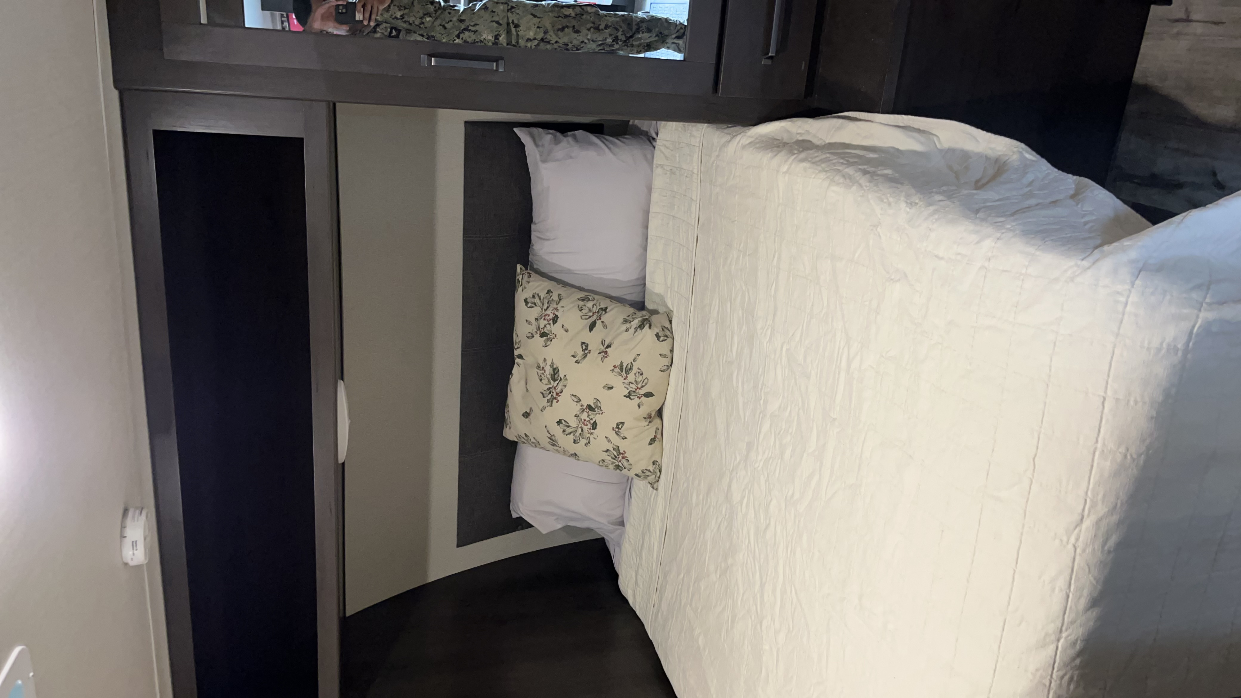 Cozy RV rental bedroom with comfortable bedding and built-in storage. Note the mirrored cabinet reflecting a person in uniform.. Grand Design Transcend Xplor 2022