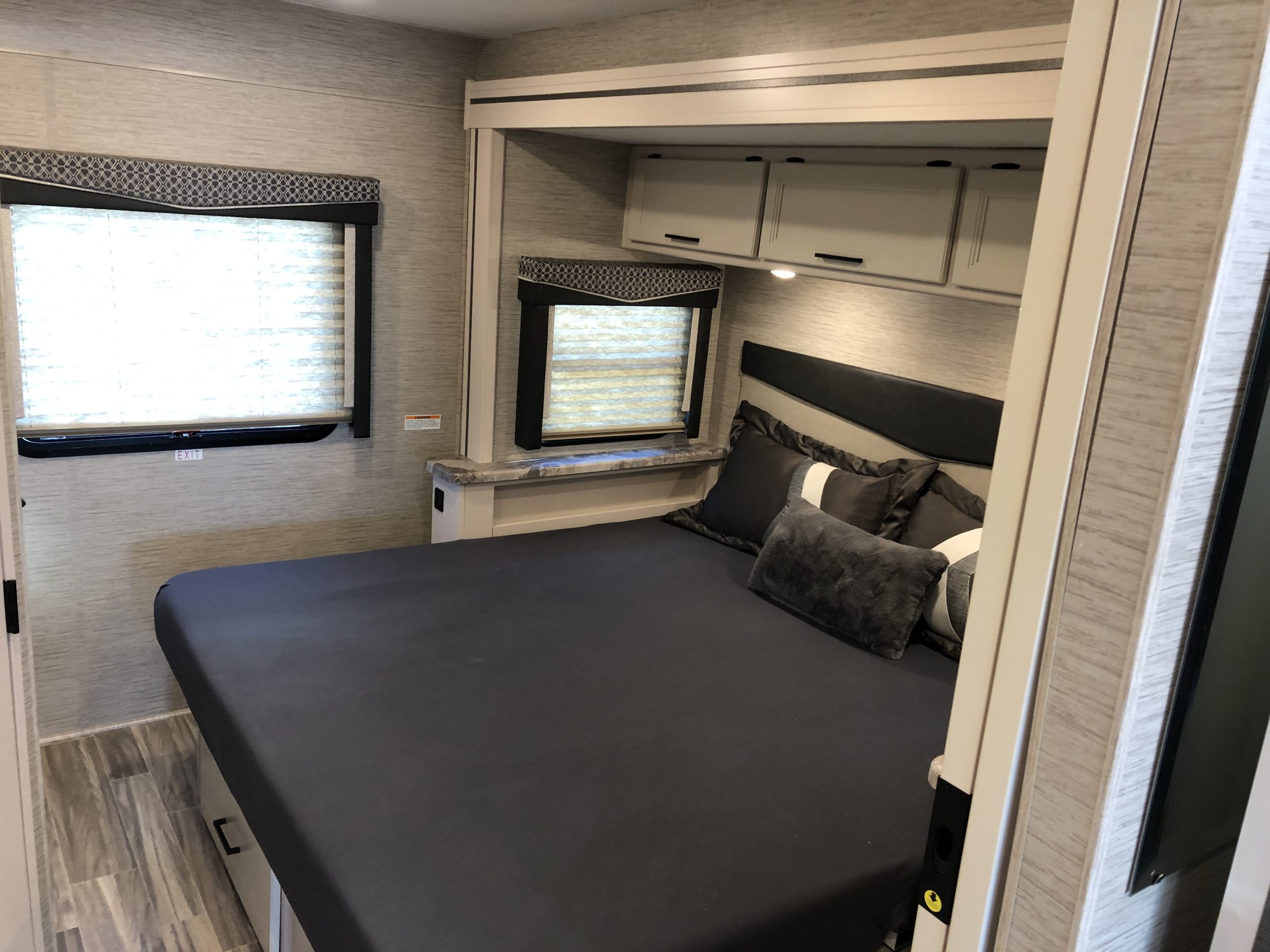 Cozy RV rental bedroom with a dark grey bed, decorative pillows, two windows with blinds, and overhead storage cabinets.. Thor ACE 2022