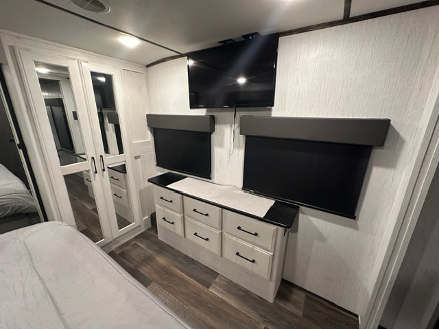 Inside an RV rental bedroom showing a wall-mounted TV, two windows with black roller blinds, a dresser, and a mirrored wardrobe.. Heartland RVs Bighorn Traveler 2022