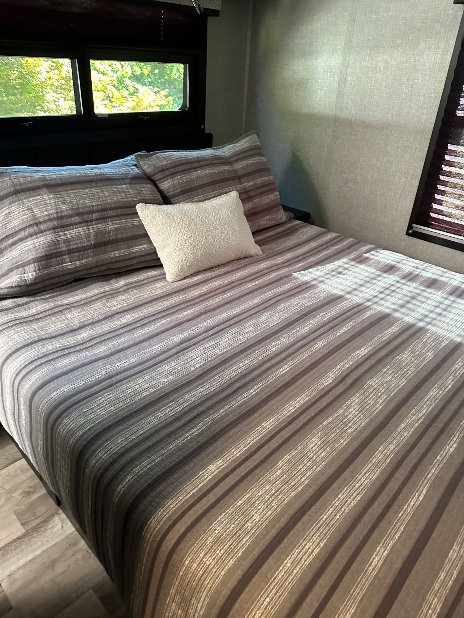 Bright RV rental bedroom: striped bed, cozy pillows, and windows show trees.. Jayco Redhawk 2023