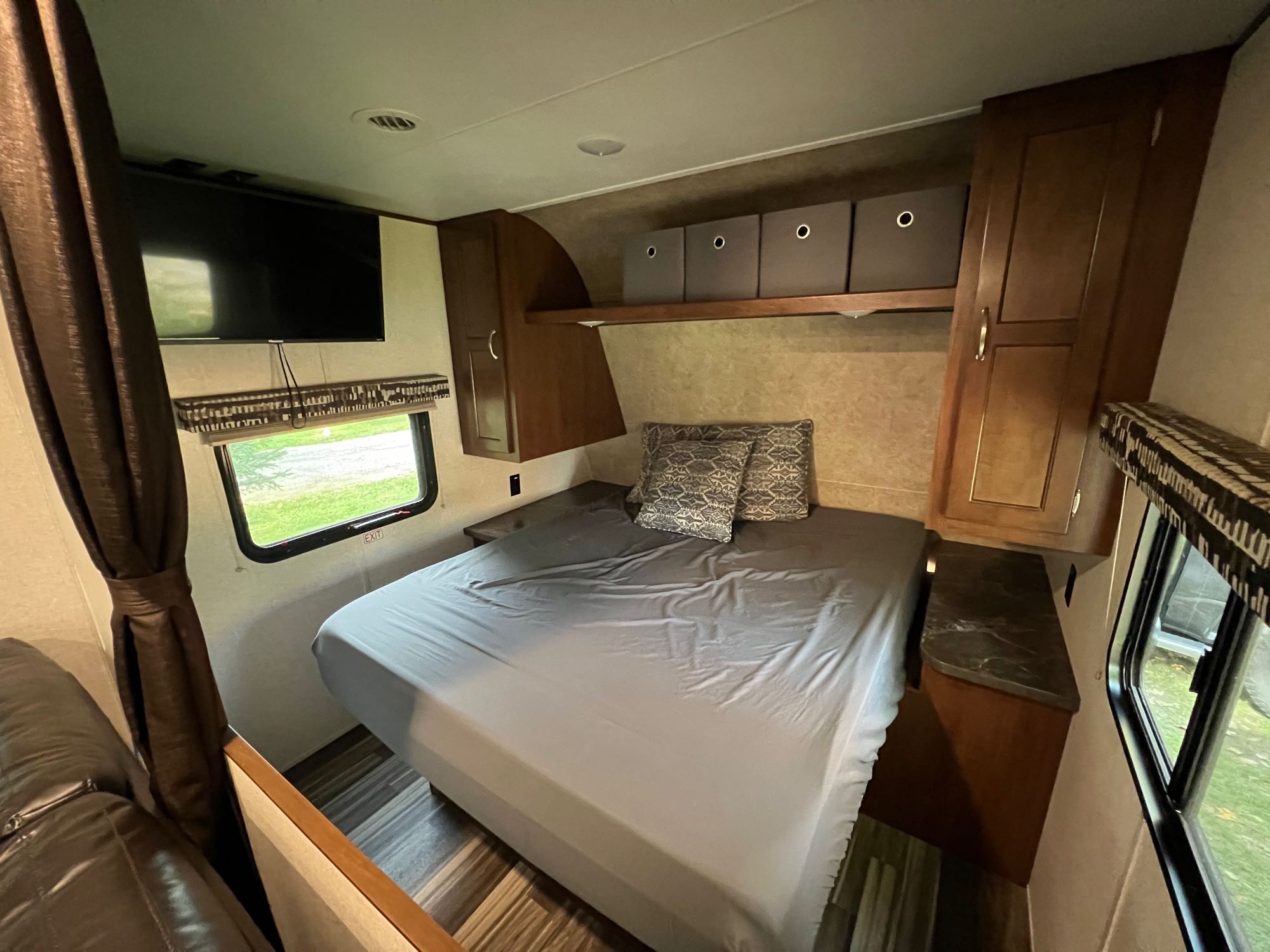This RV rental shows a comfy bedroom with a gray-sheeted bed, wall-mounted TV, overhead storage bins, and wood-look flooring.. Jayco Jay Flight SLX 2017
