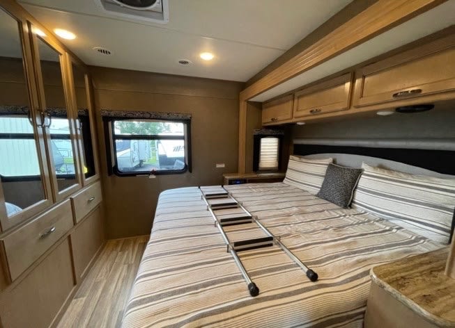 Spacious RV rental bedroom featuring a striped bed with ladder, light wood cabinets, and two large windows.. Thor A.C.E. 29.4 2018