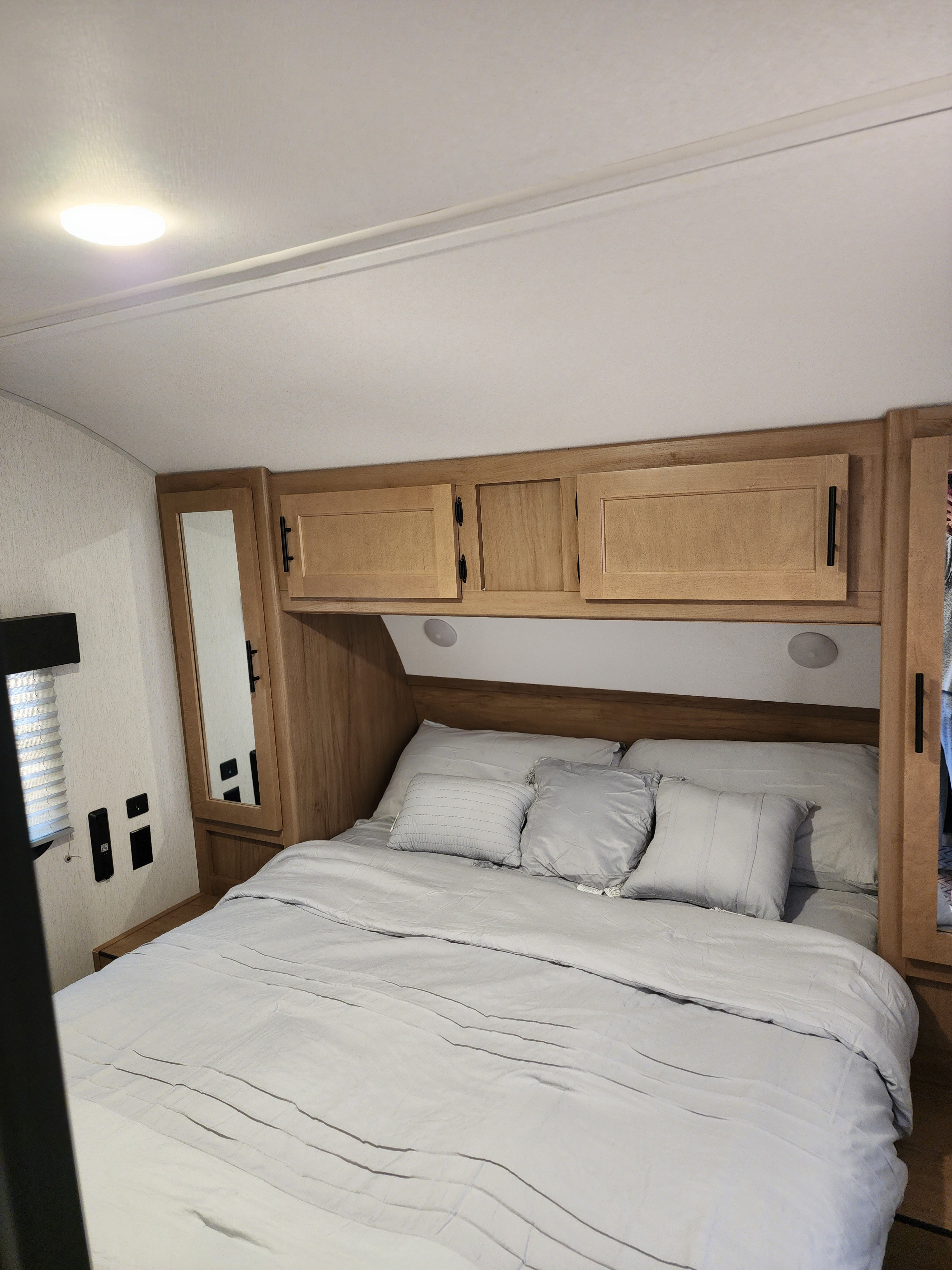 This RV rental features a cozy bedroom with a comfortable bed, light gray bedding, wood overhead cabinets, and a side mirror.. Forest River Cherokee 2023