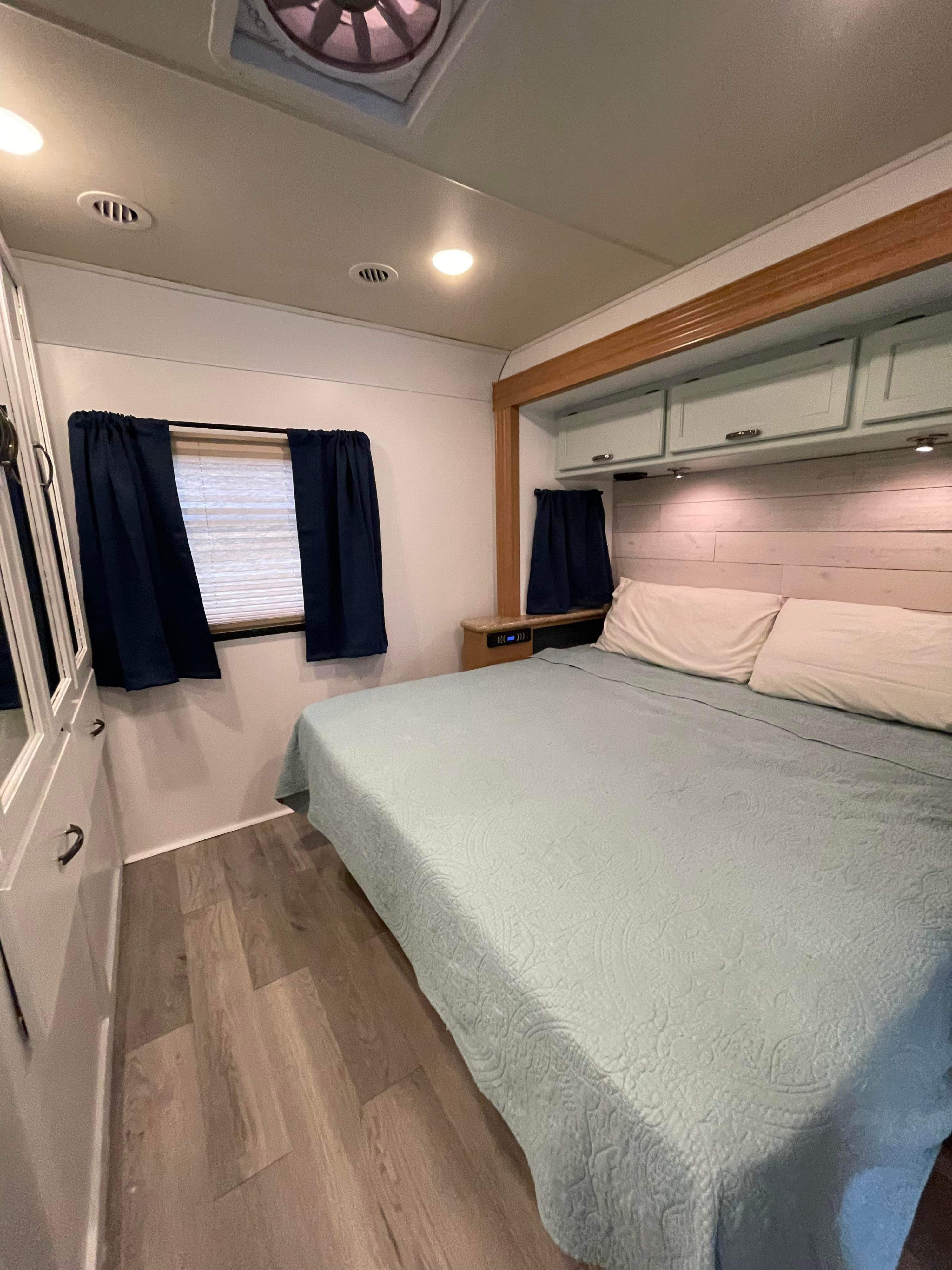 RV rental bedroom: light blue quilted bed, white pillows, overhead storage, blue curtains, and wood-look floor.. Thor A.C.E. 2016