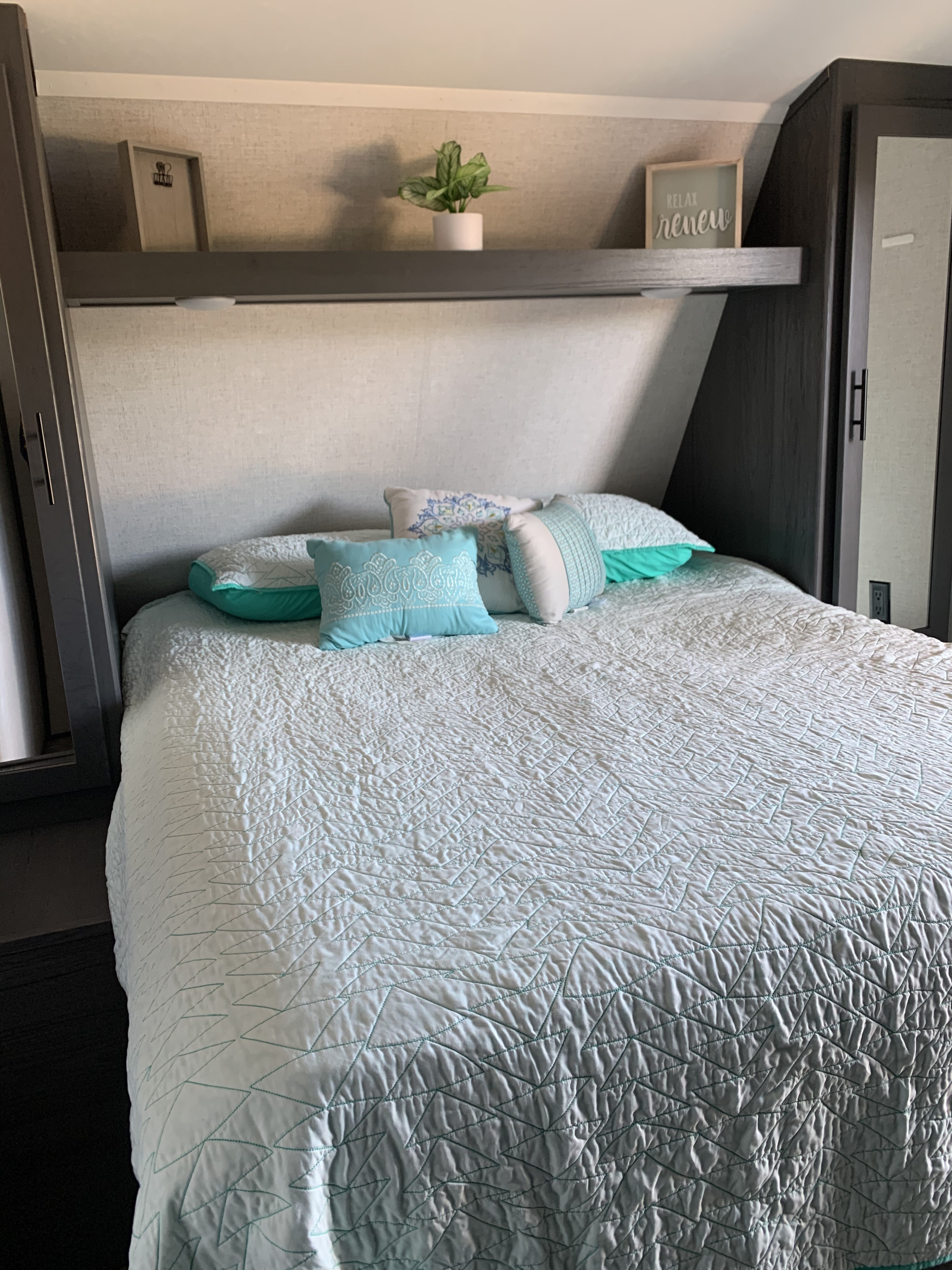 Cozy RV rental bedroom with a light blue quilted bed, teal pillows, and decorative items on a shelf, perfect for relaxing.. Dutchmen Other 2021