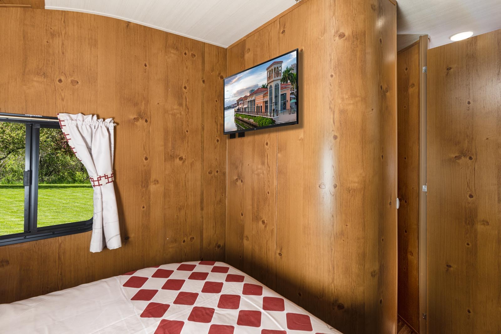 This RV rental offers a cozy wood-paneled bedroom with a comfy bed, TV, and a window providing a green outdoor view.. Gulf Stream Cruiser 2021